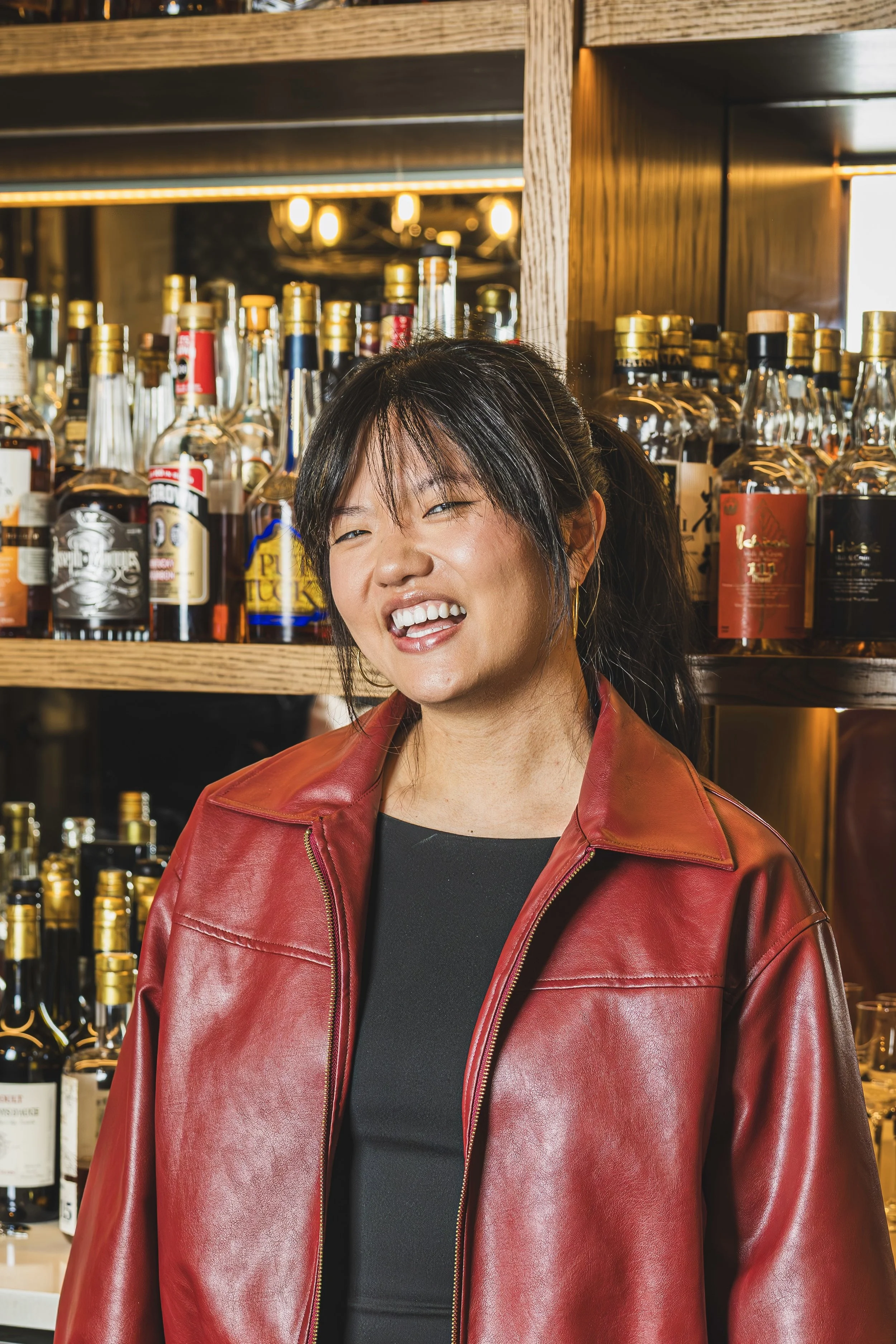 A woman standing behind a bar counter with various bottles and cans, smiling and looking to her left, in a bar or restaurant setting with a rustic brick wall background.