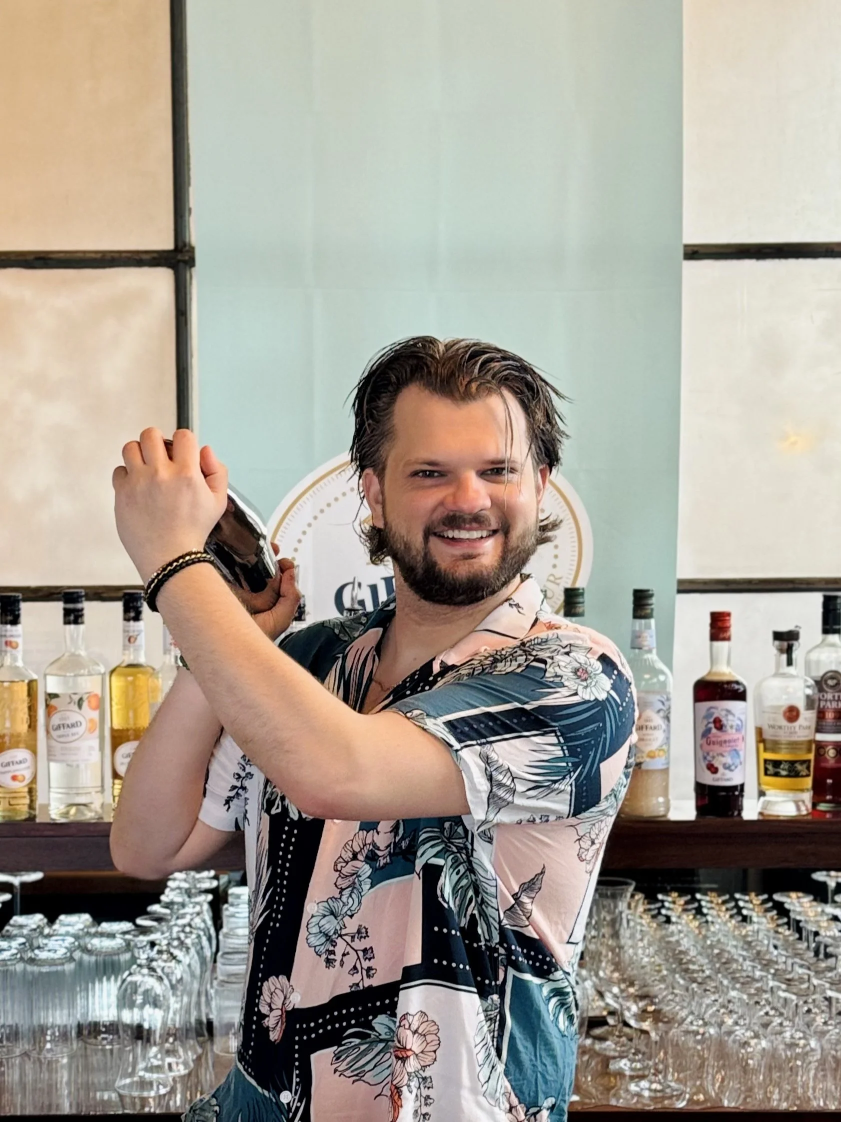 A smiling man with dark hair and a beard, wearing a black button-up shirt, standing behind a bar with plastic cups and a rubber mat.