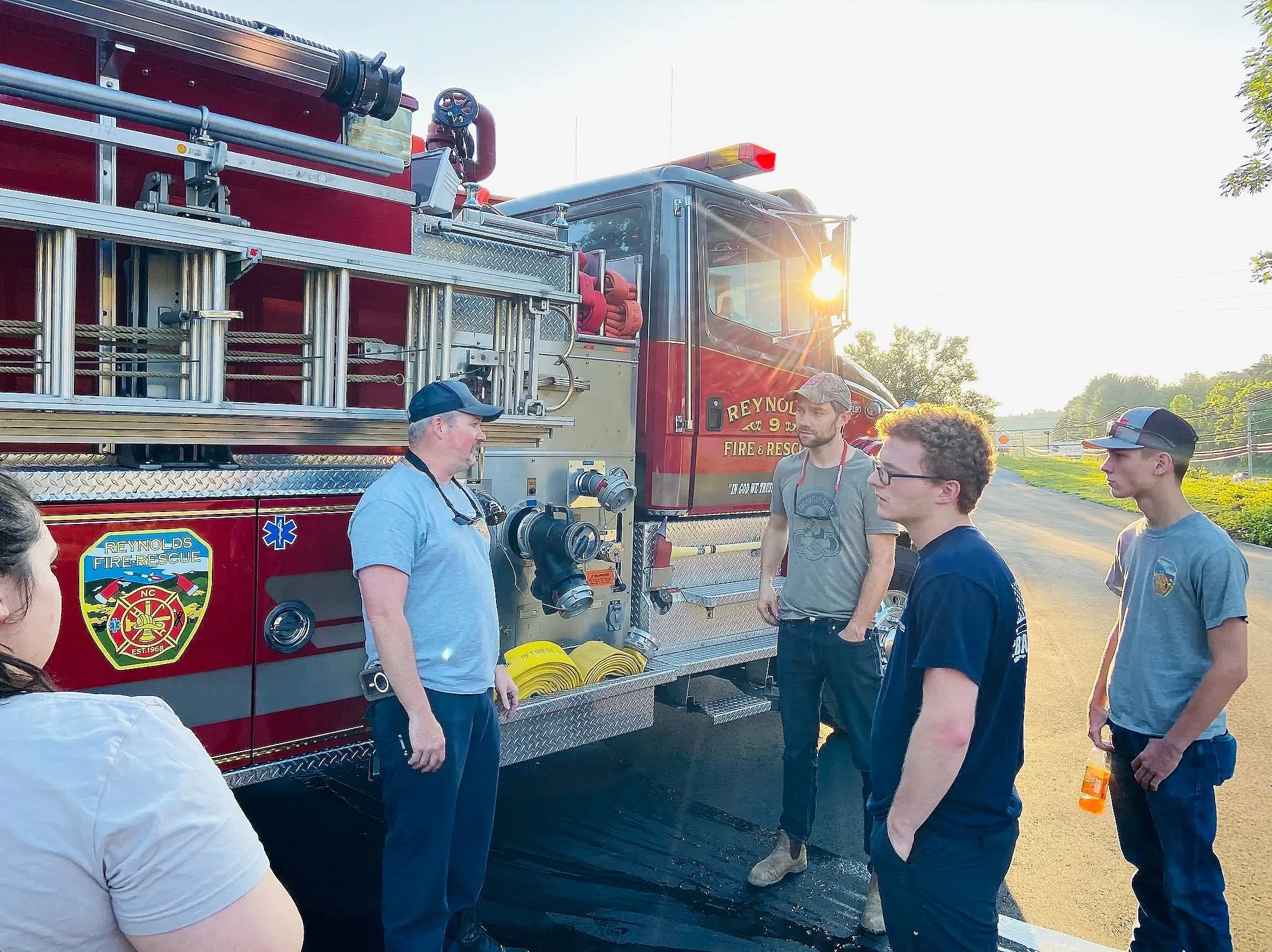 Firefighters gathered around a red fire rescue truck on a rural road at sunset. The truck has fire hoses and equipment on it, with badge visible. Sunlight shines through the truck's window as the group listens to a firefighter.