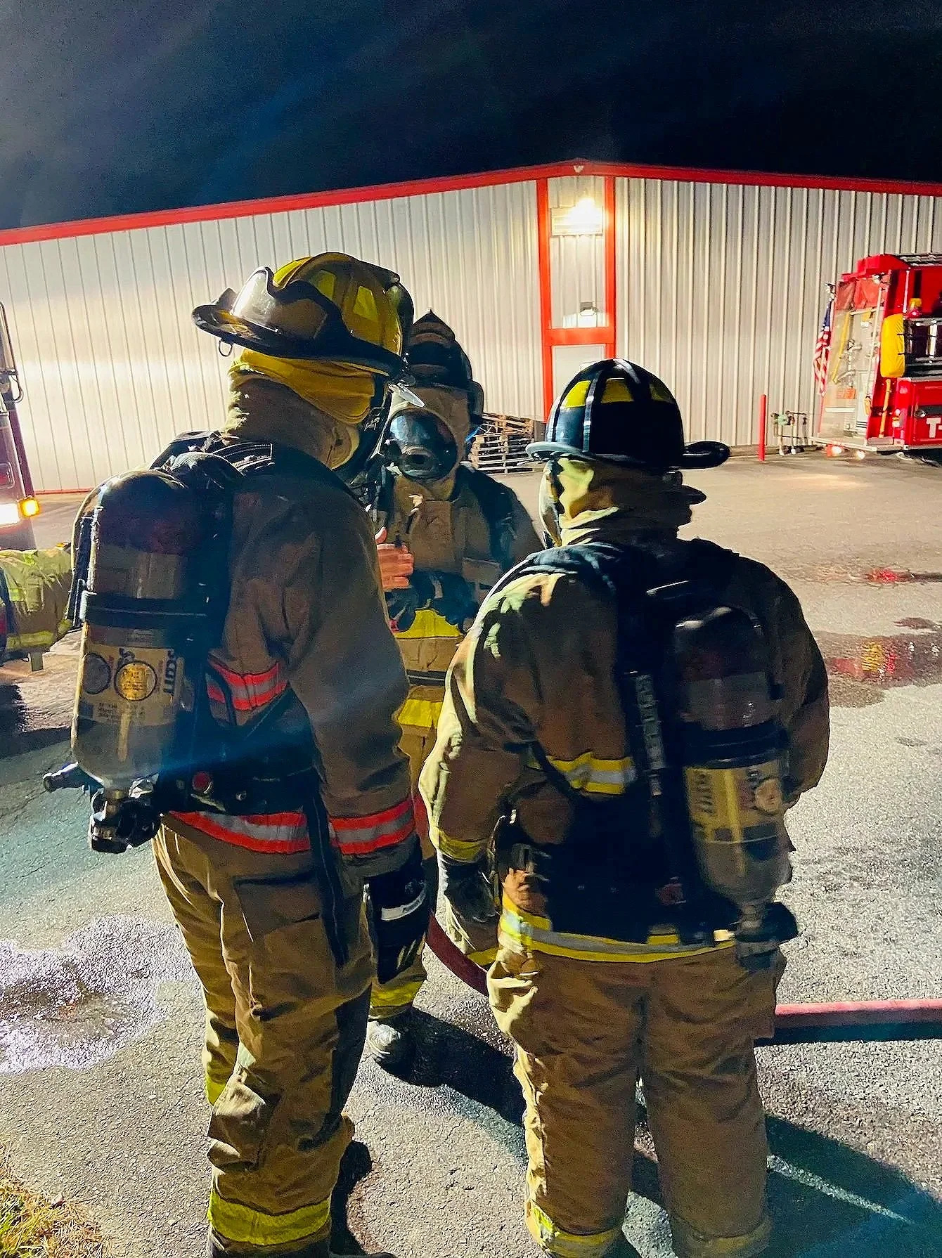 Drew and fellow firefighters in full gear standing outside at night near a fire station, engaging in conversation.