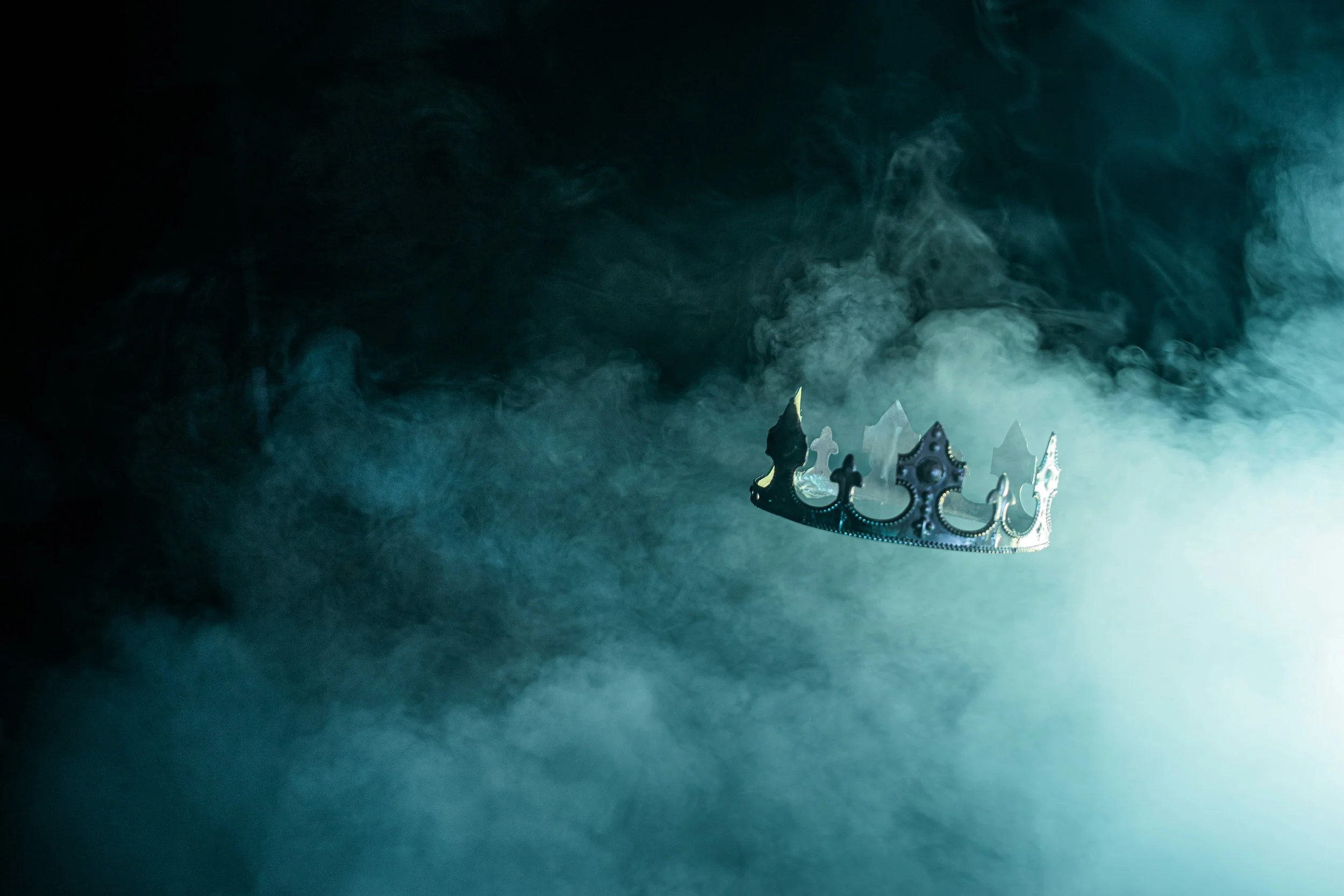 A silver crown floating amidst blue and white smoke with a dark background.