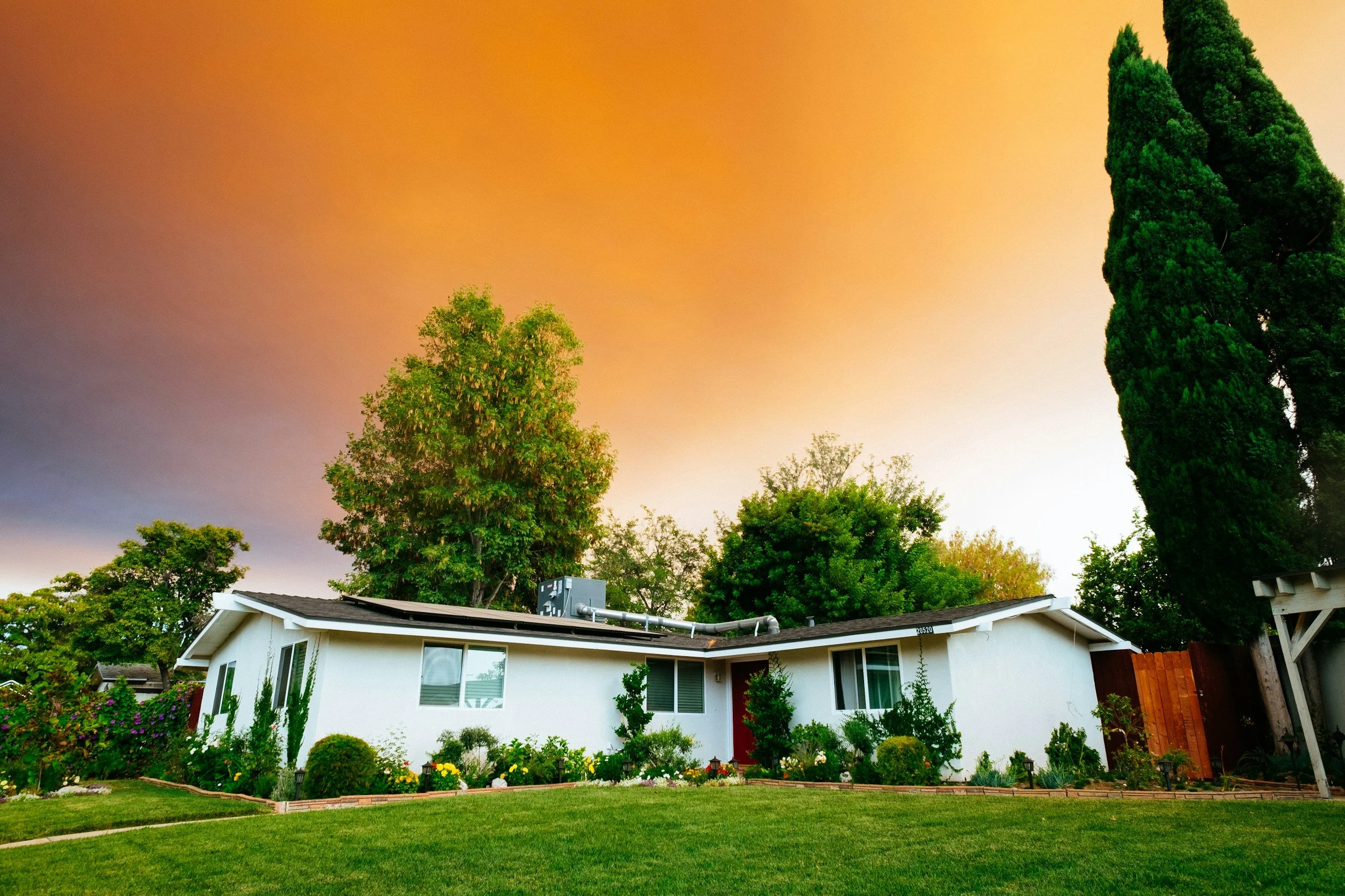 A white house with a landscaped front yard and trees, under a colorful sunset sky.