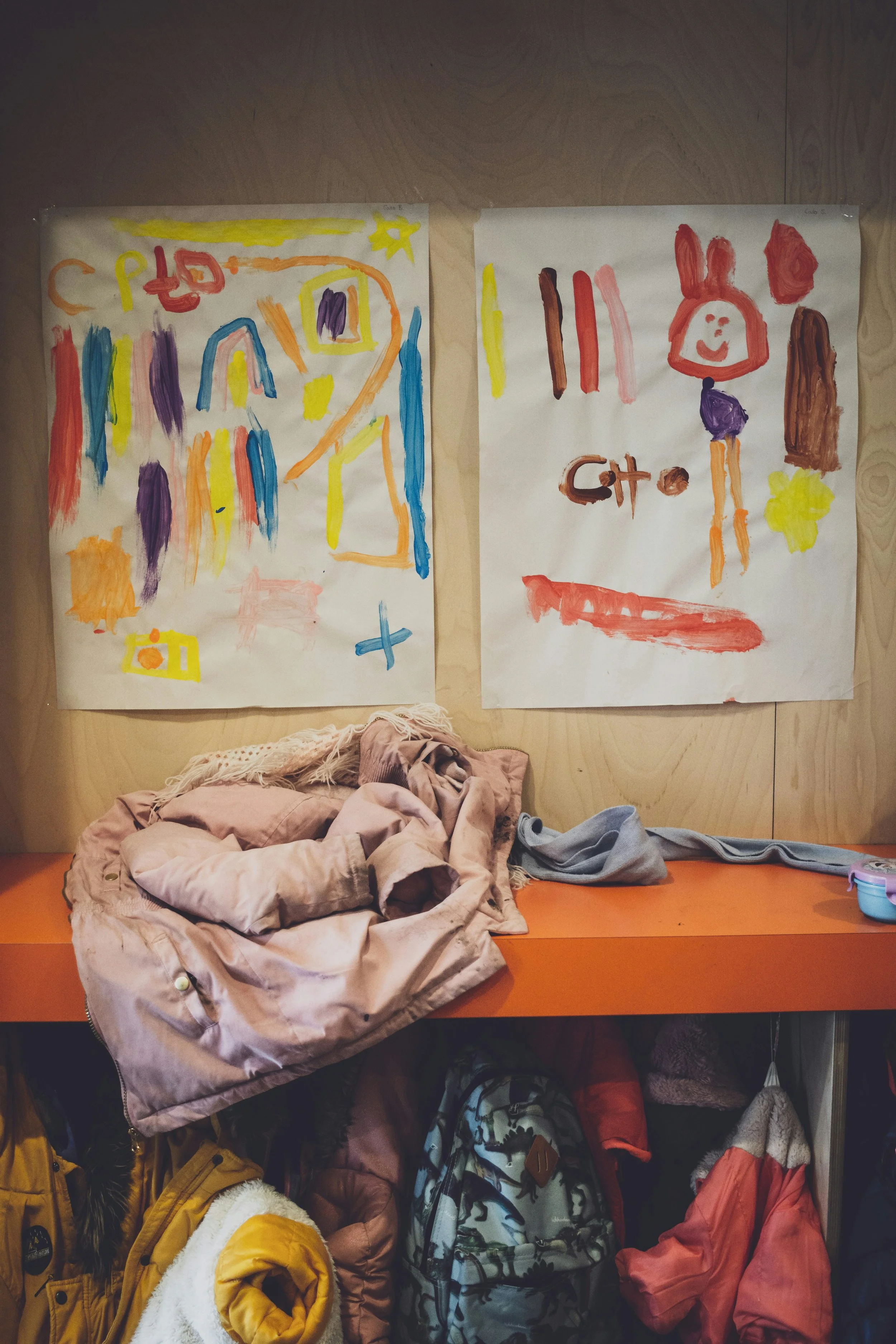 Two large colorful children's drawings on paper are hung on a wooden wall, with a pile of pink and gray jackets and backpacks underneath.