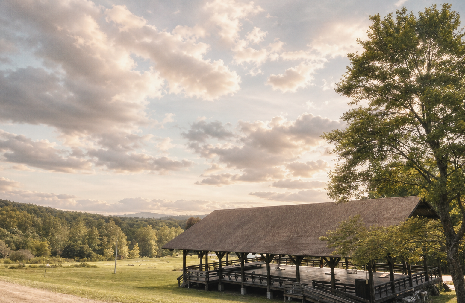 A rustic pavilion with a brown roof situated in a green landscape under a partly cloudy sky.