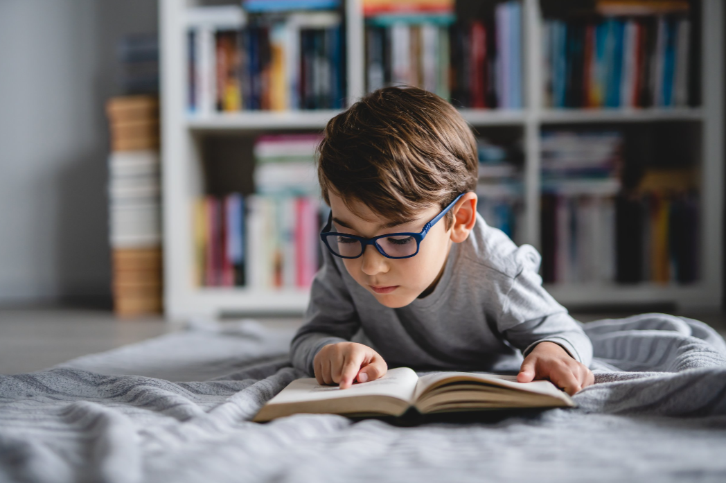 A young boy wearing glasses lays on a blanket reading a chapter book