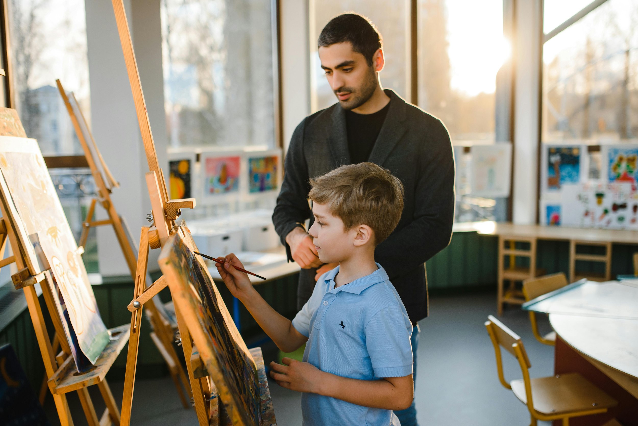 A student artist paints a picture while his tutor stands nearby
