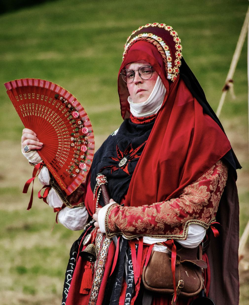 A woman outside in a backyard with trees, wearing glasses and a costume headpiece resembling a historical or fantasy character, with feathers and fabric in red, white, and black.