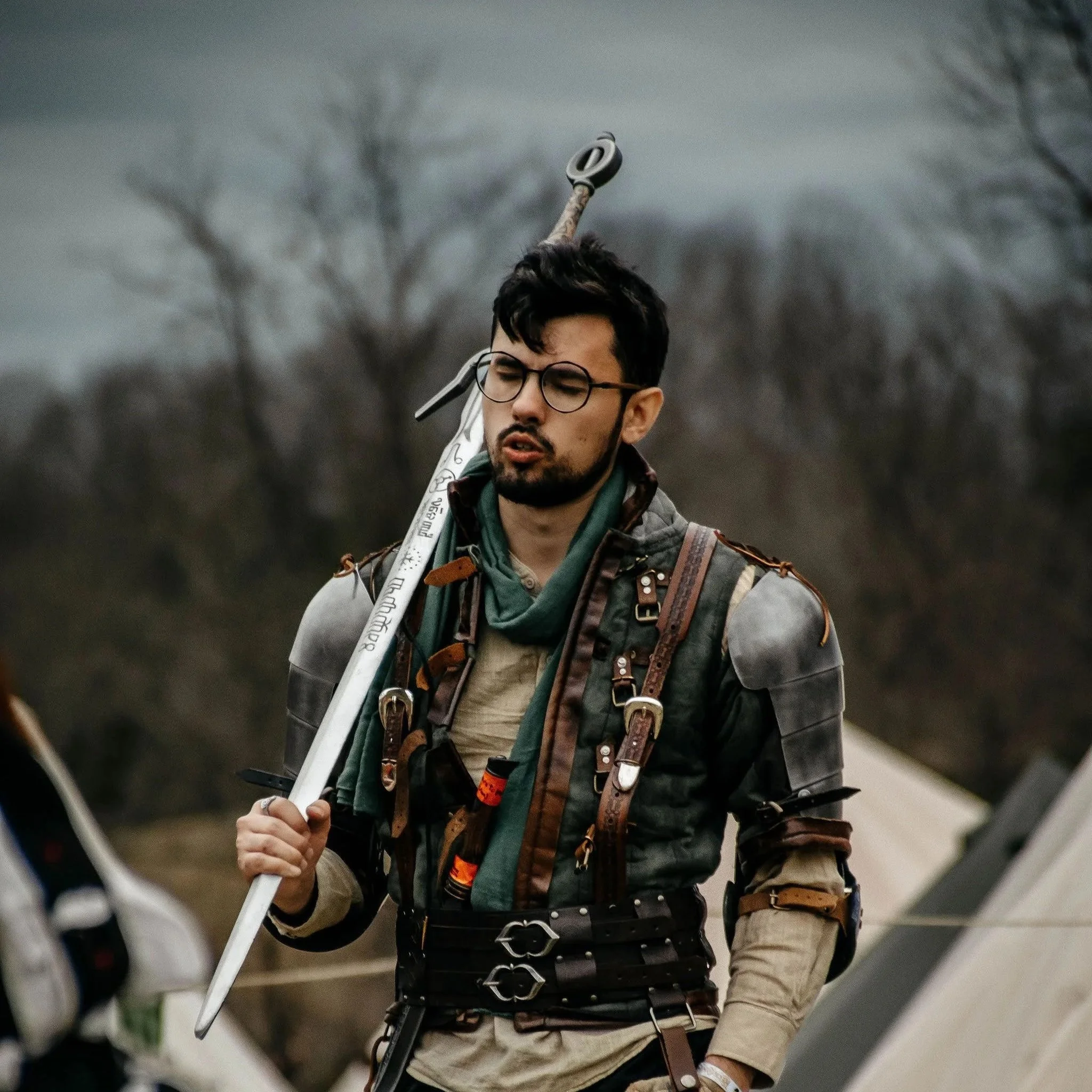 A man with glasses and dark hair, dressed in a fantasy or medieval costume, carrying a large sword on his shoulder, outdoors with trees and tents in the background.