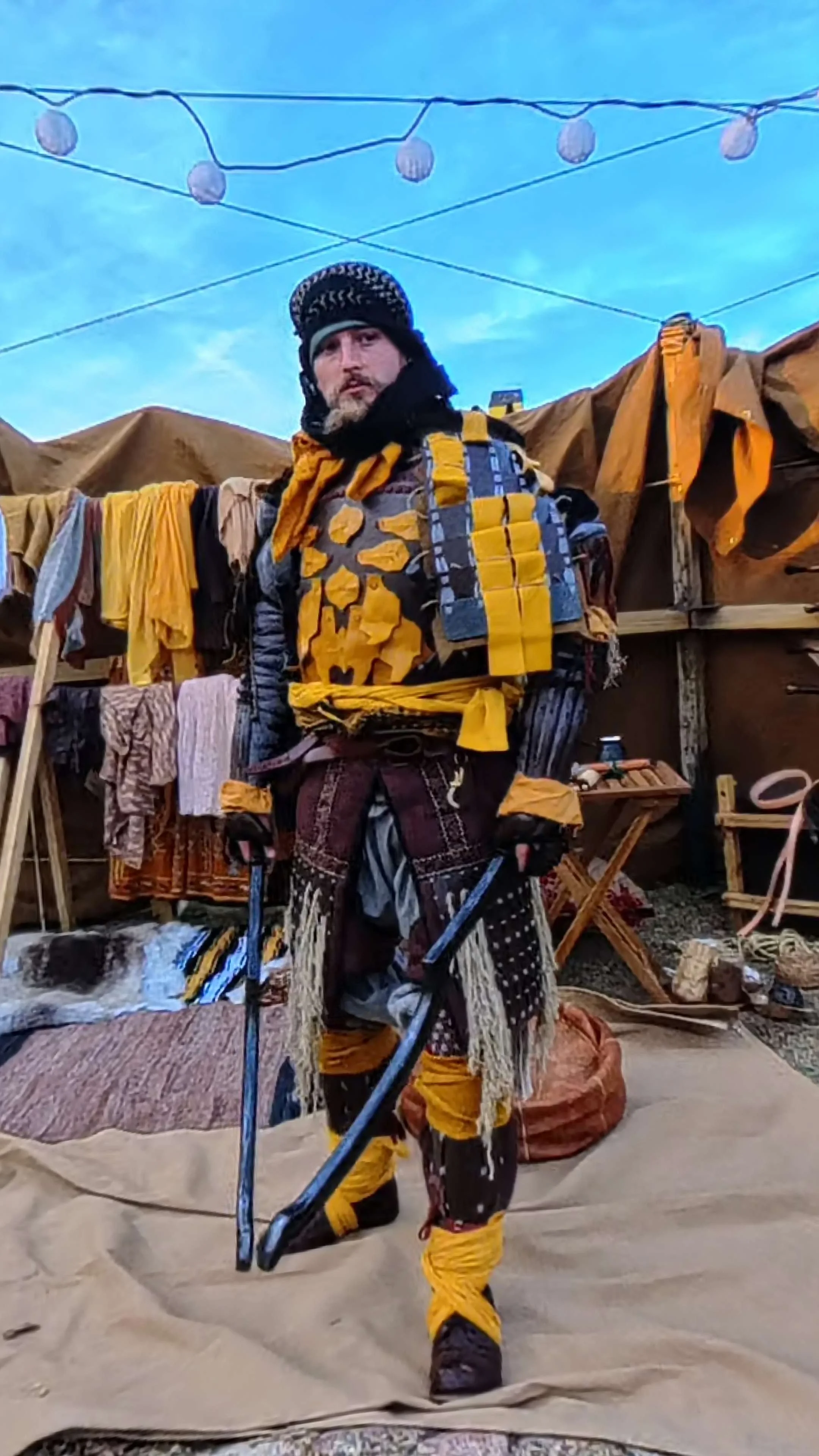 A woman dressed as a medieval or fantasy character, wearing a gold-colored robe with embroidered blue and gold details, a large ornate pendant necklace, red earrings, and a dragonfly brooch, standing outdoors in a wooded area with trees in the background, holding a sheathed sword.
