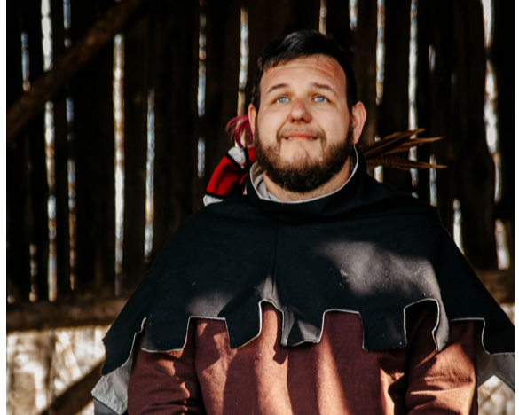 A man with a beard and short hair, wearing a black cape and a red and black backpack, standing in front of a wooden forest background.
