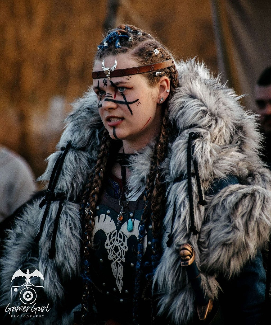 A young man with face paint resembling tribal or warrior markings, wearing a leather necklace with beads and a decorative shoulder piece, standing outdoors among a group of people.