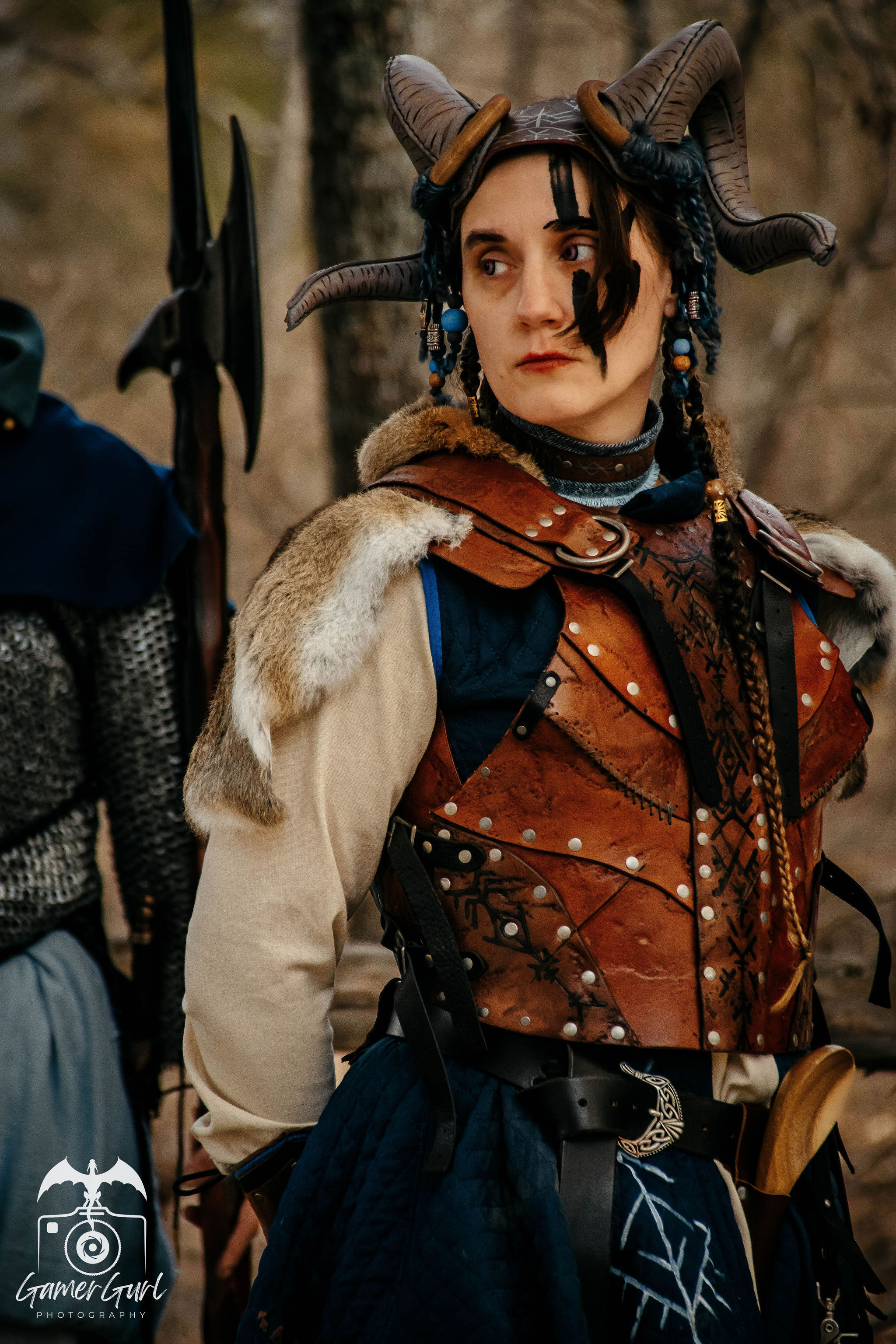 A man dressed as a medieval or fantasy character wearing a blue tunic with a hood, layered necklaces with charms, and a headband, holding a small sword or dagger in a leather sheath at his waist, standing outdoors with a tent in the background.