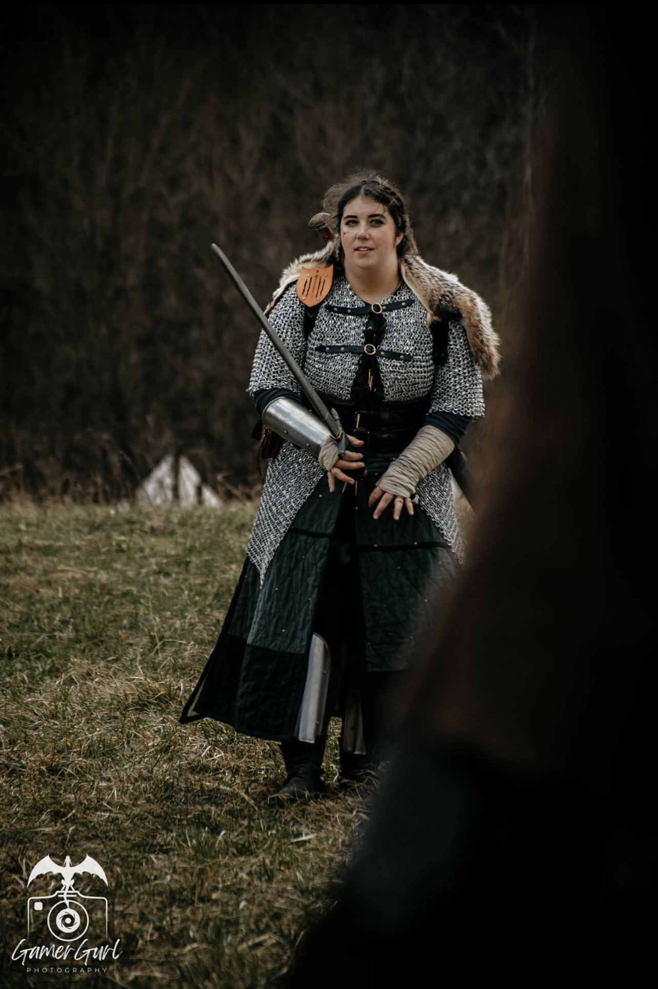 Middle-aged woman with gray hair pulled back, wearing a green dress with a decorative necklace, holding a mug, standing among a group of people outdoors.
