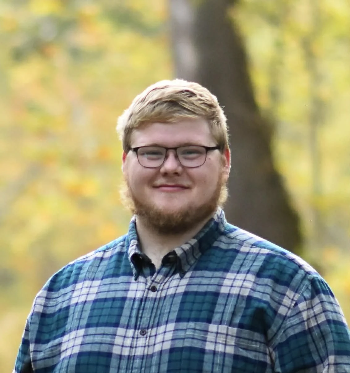 A young man with glasses, a beard, and blonde hair, smiling outdoors with autumn trees in the background.