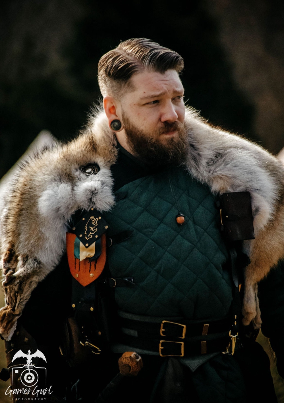 A woman with brown hair, glasses, and a purple headband standing outdoors among trees, dressed in period costume with a green cape, black dress, and leather belt holding leather pouches.
