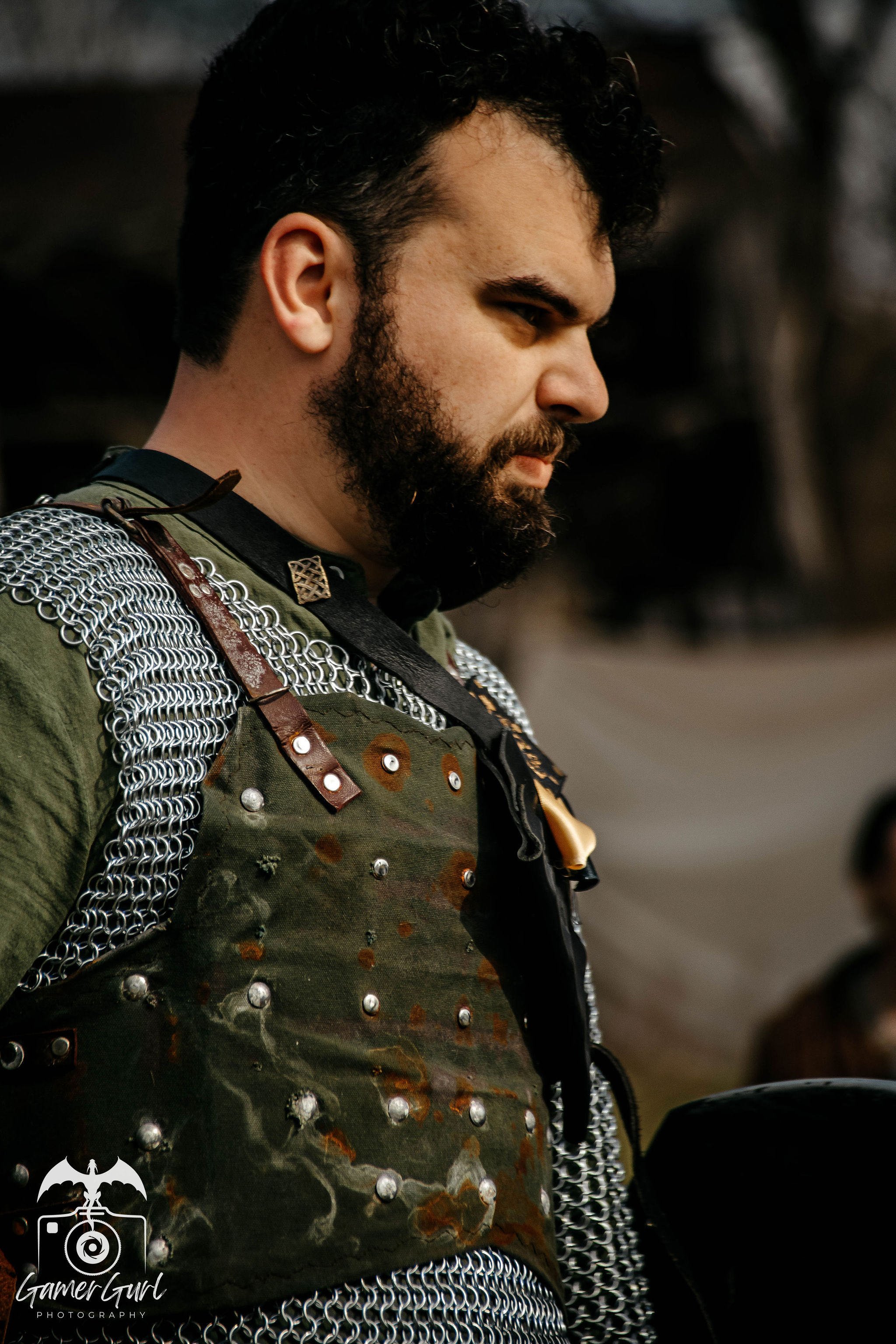 A man with dark, curly hair and a beard wearing medieval-style clothing, standing among others dressed similarly, in an outdoor setting.