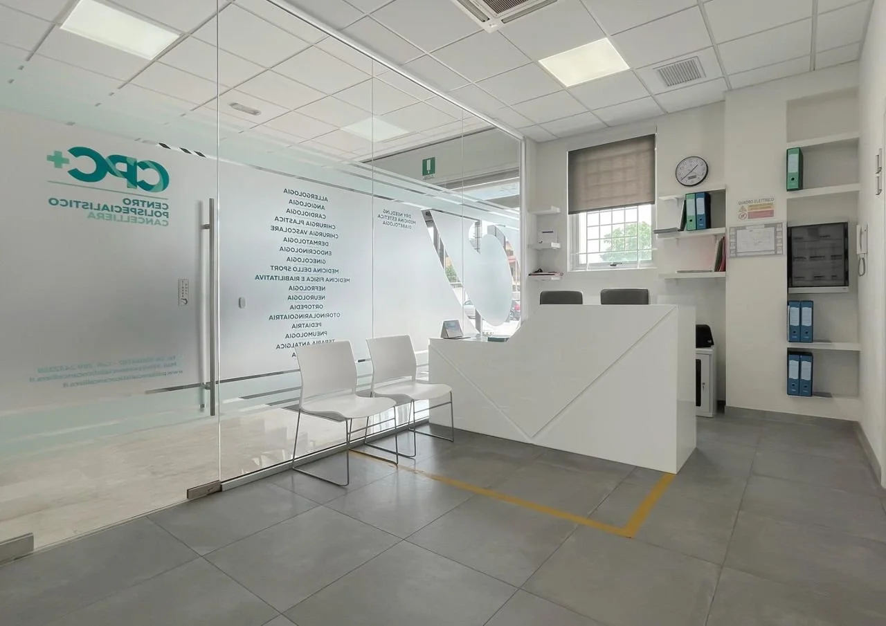 Reception di uno studio medico con sedie e scrivania, pareti con scaffali e una finestra.