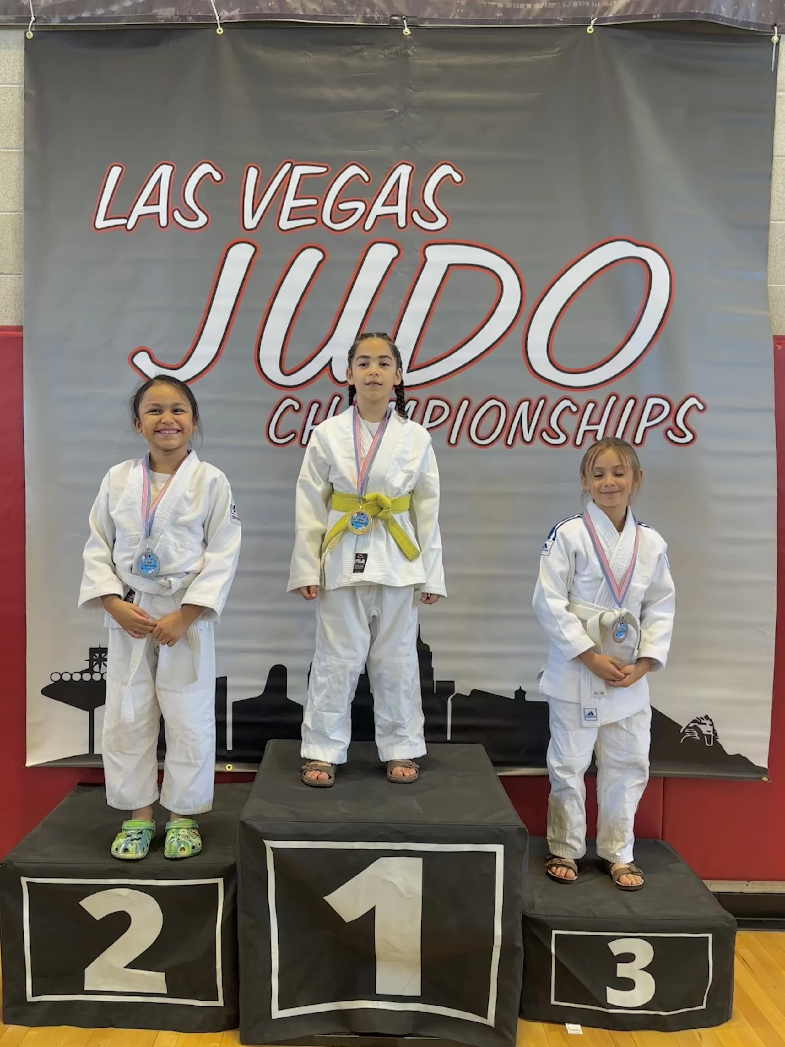 Three young girls in judo uniforms standing on winners' podium at a judo championships, with medals around their necks, in front of a banner that reads 'Las Vegas Judo Championships'.