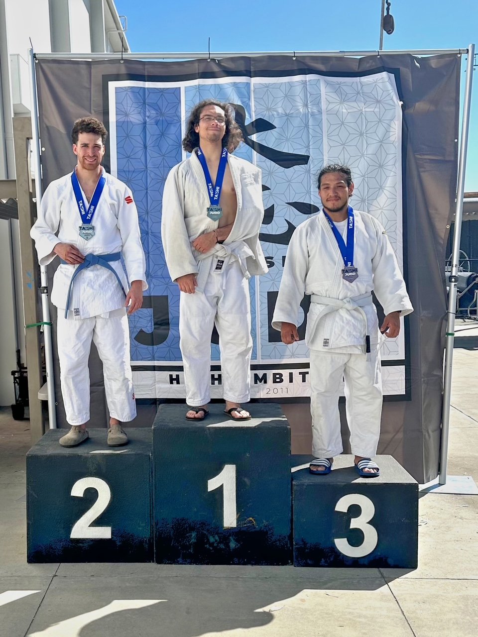 Three male judo practitioners standing on a winners' podium with medals around their necks, wearing judo gis. The person in the center, on the first-place platform, is on top with a confident expression, while the second-place and third-place winners
