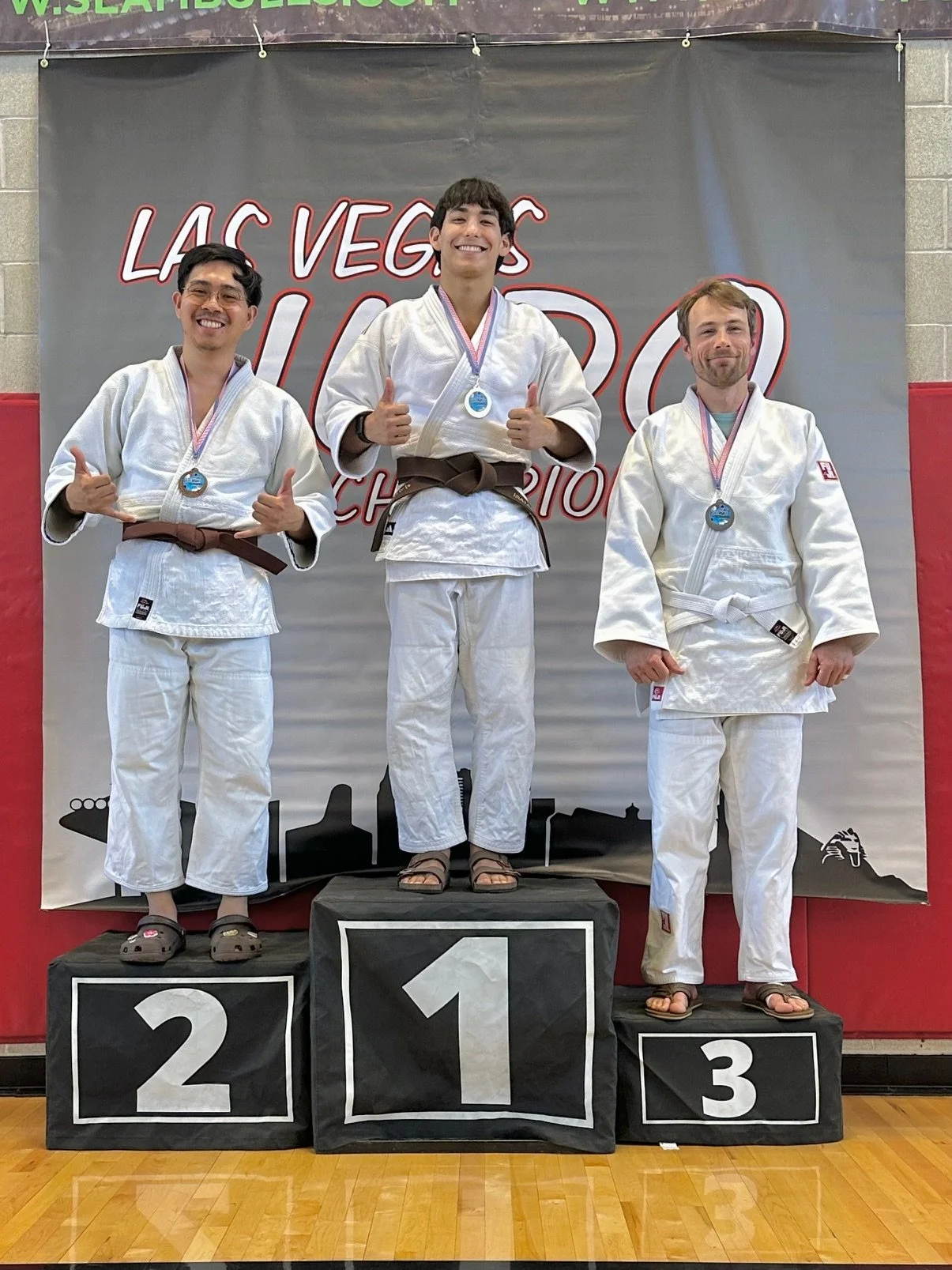 Three judo athletes on a winners' podium. The first-place winner in the center has a brown belt and is giving a thumbs-up. The second-place athlete on the left has a brown belt and is making a shaka sign. The third-place athlete on the right has a wh
