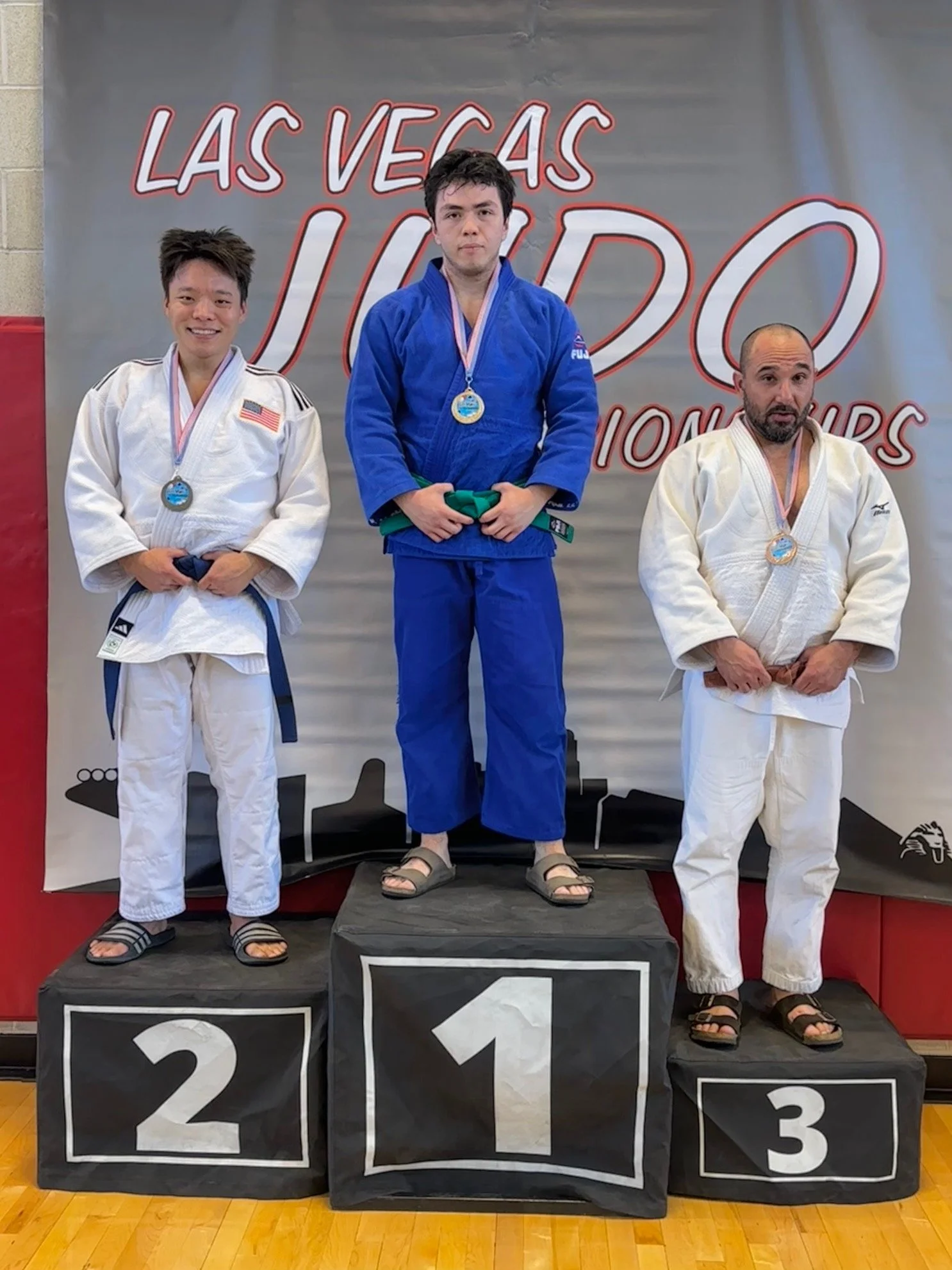 Three male judo athletes on a winners' podium at a competition, wearing medals and judo gi uniforms, with the first-place winner in the center on the highest step, second to the left, and third to the right.