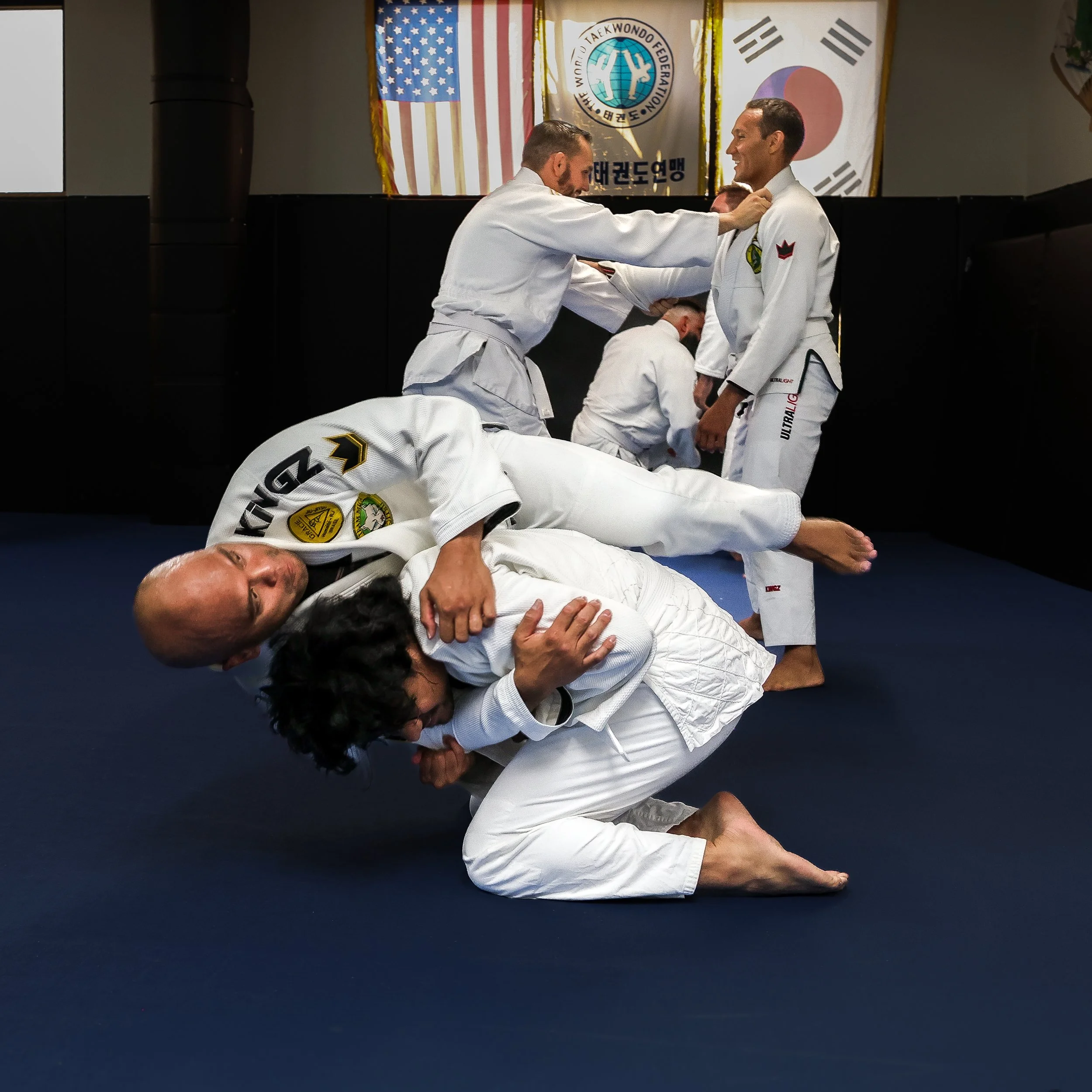 Two women practicing judo; one woman is lying on the mat with her arm extended, and the other woman is kneeling beside her, holding her arm.