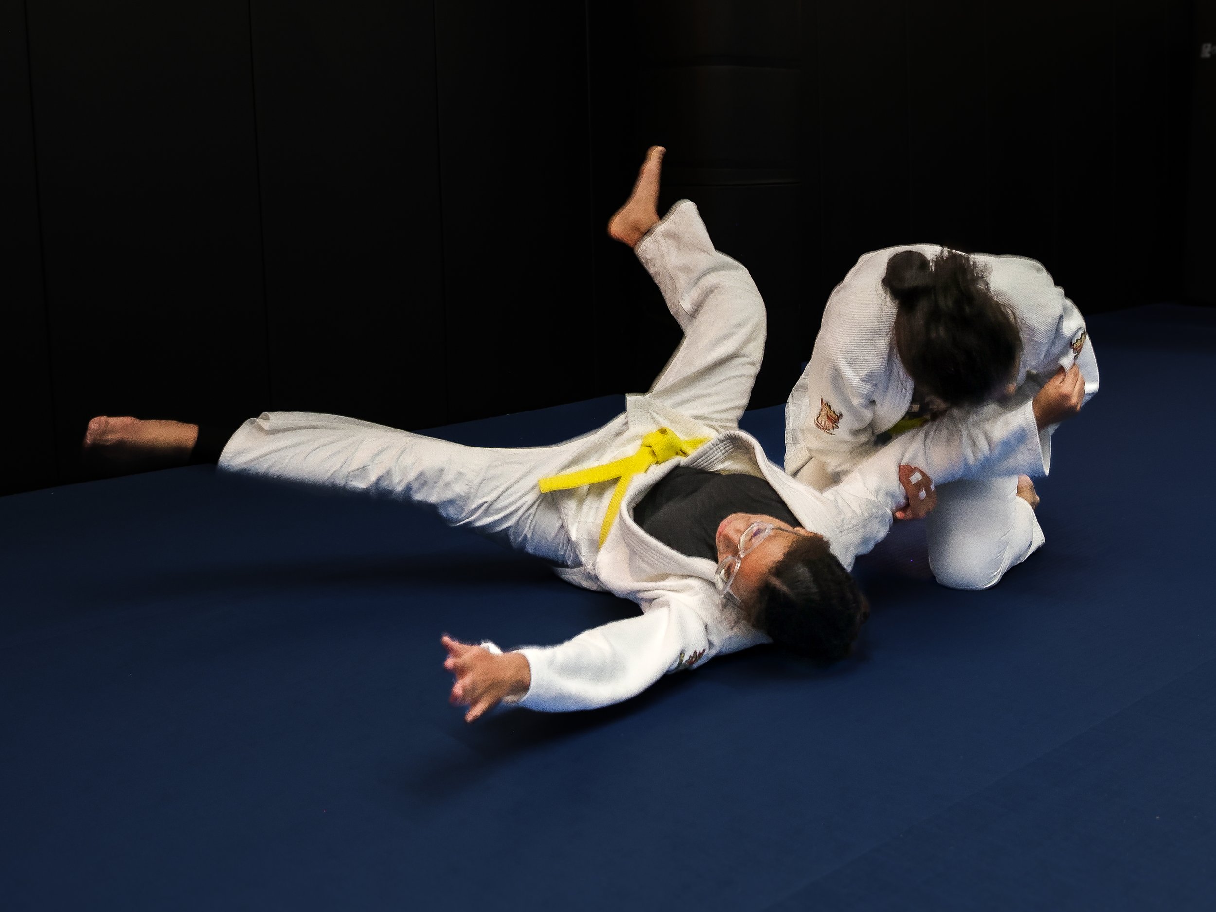 Two women practicing judo on a blue mat, with one woman on the ground and the other on top, both wearing martial arts gi uniforms.