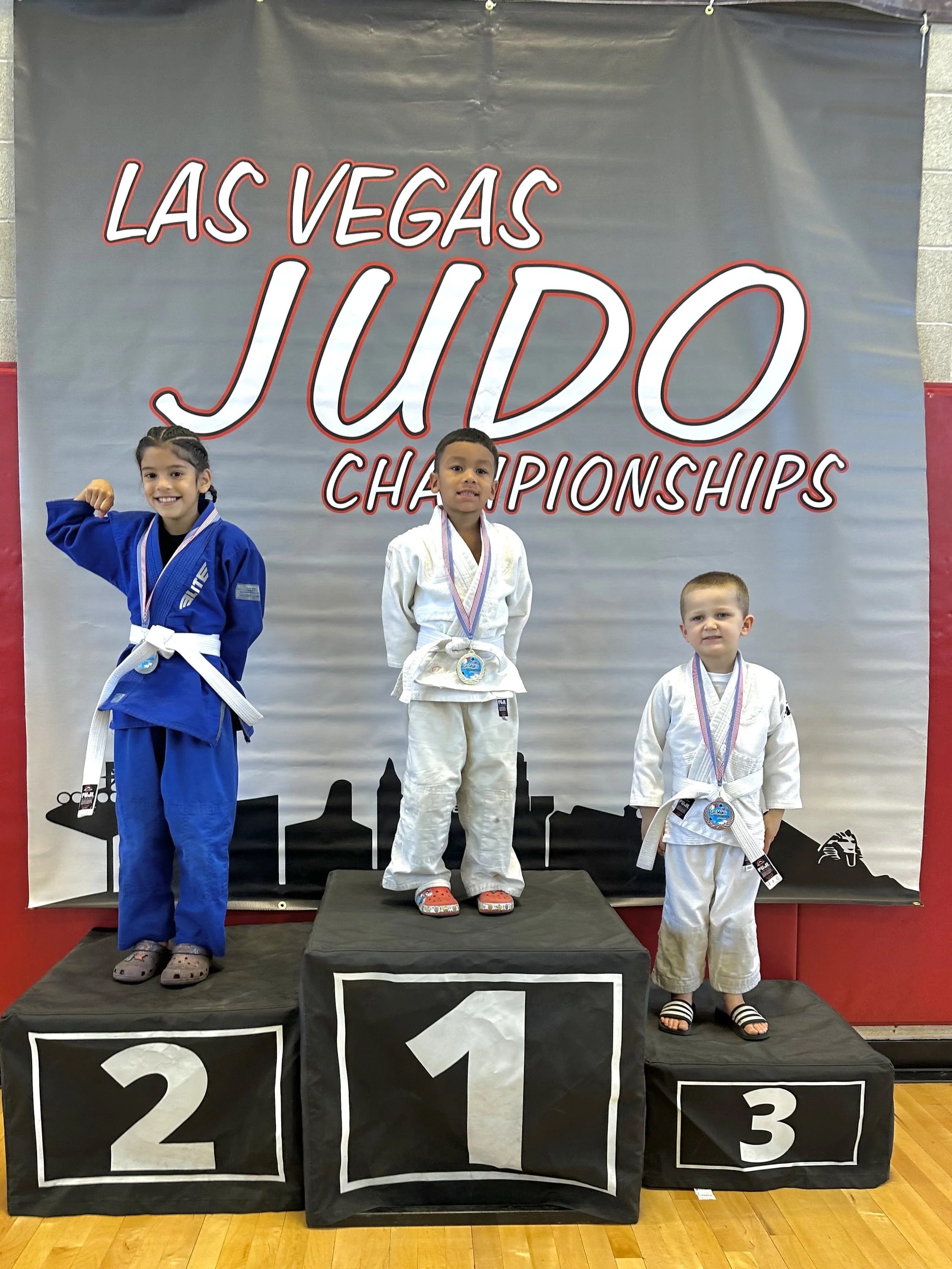 Three young children standing on winners' podium at Las Vegas Judo Championships, wearing judo gis with medals.