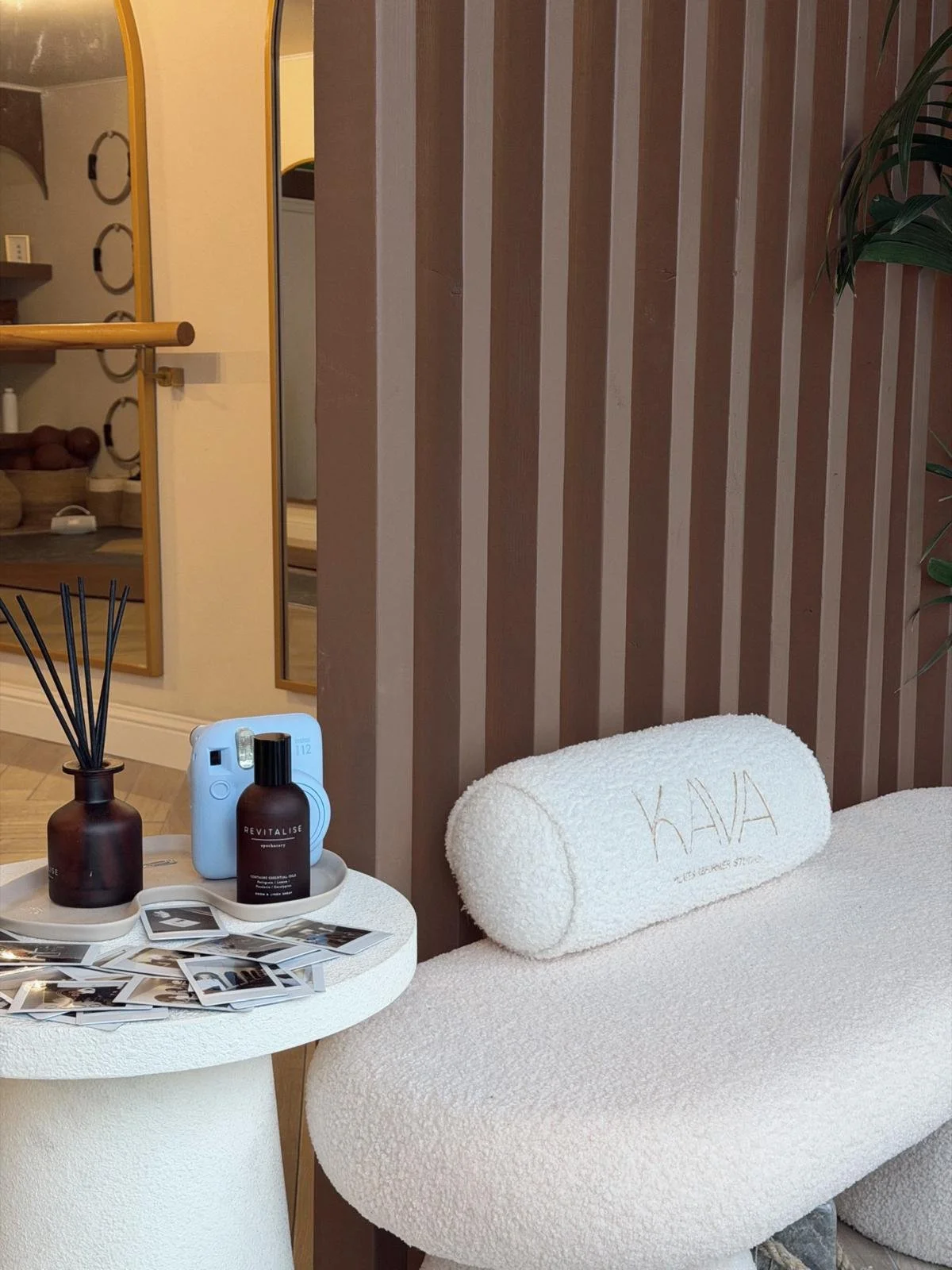 A cozy interiorwith a white, textured bench that has a  embroidered with the word on it, a round white side table holding a dark brown diffuser with reed sticks, a small brown bottle labeled "Revitalise," a white portable speaker, and scattered polaroid-style photographs. There's a wooden slatted wall behind the bench