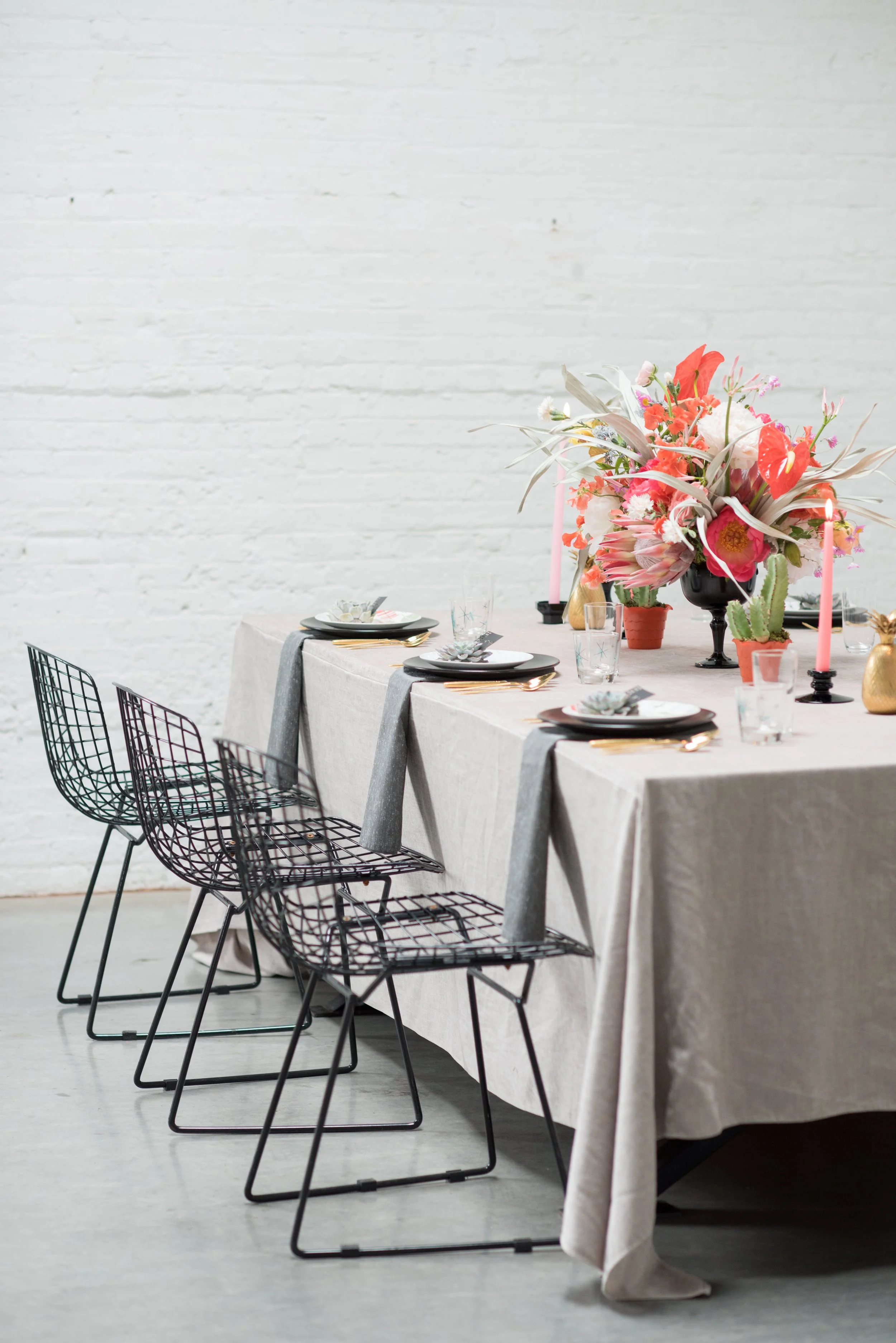 A row of three place settings are on a table with a cream tablecloth, black wire chairs, and tropical floral arrangements.