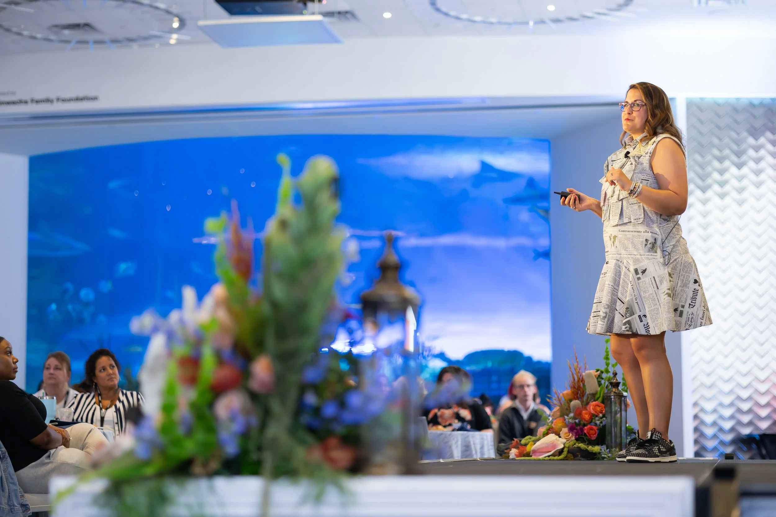 Leah Weinberg, lawyer and speaker, stands on a stage wearing a black and white newspaper patterned dress. In the background is a shark-filled aquarium and guests watching Leah speak.