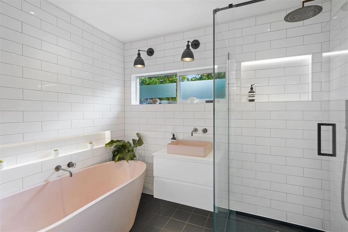 Modern bathroom with white subway tiles, black fixtures, a pink bathtub, a walk-in shower with glass door, a pink sink, and a window with frosted glass and greenery outside.