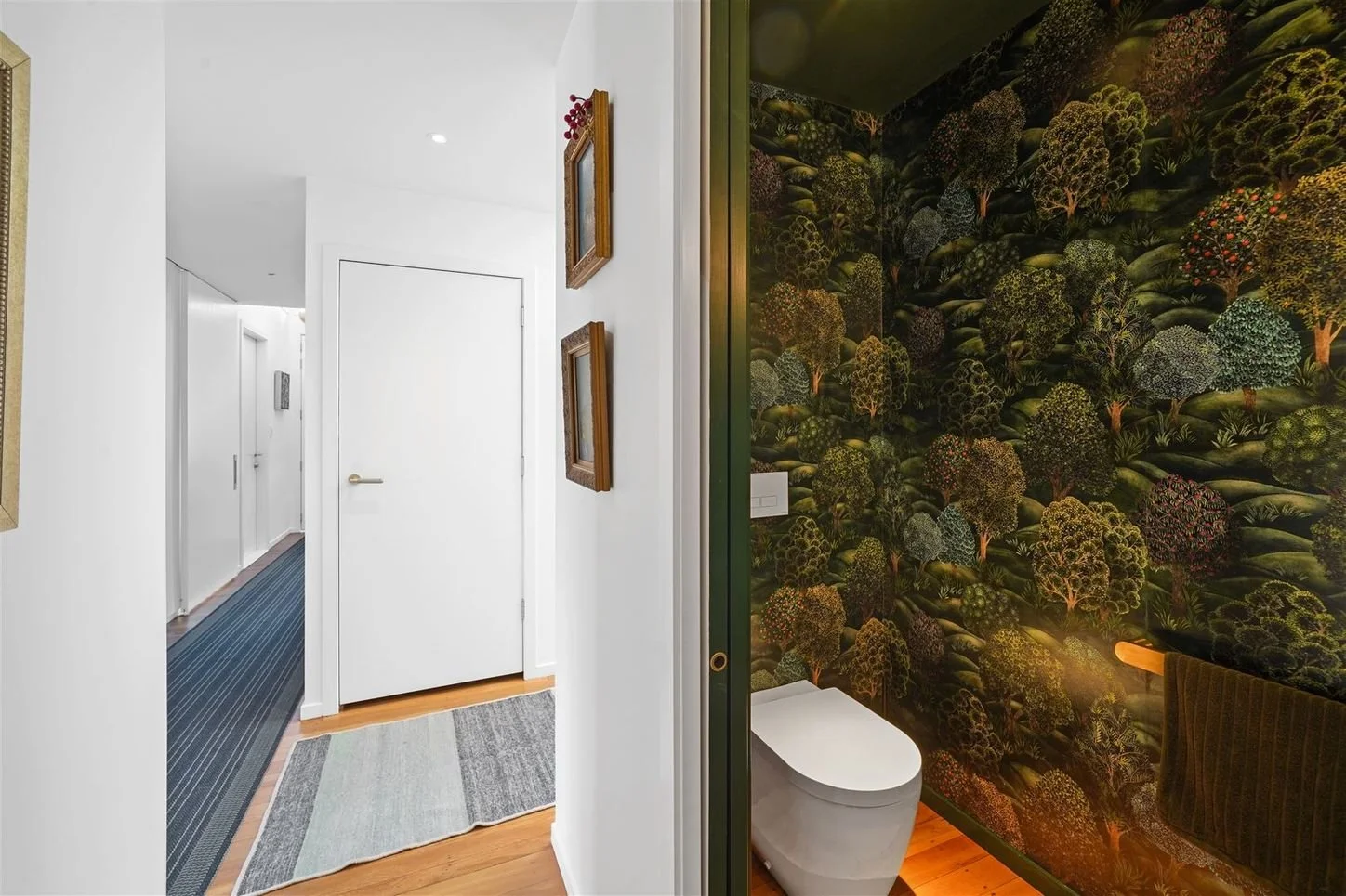 Interior view showing a corridor with white walls, closed white doors, and a bathroom with a forest-themed wallpaper and a modern toilet.