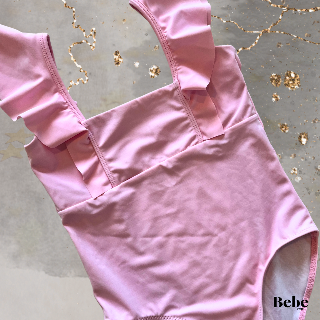 Pink girls one-piece swimsuit with ruffled straps, laid on a grey surface with gold and star-shaped confetti.