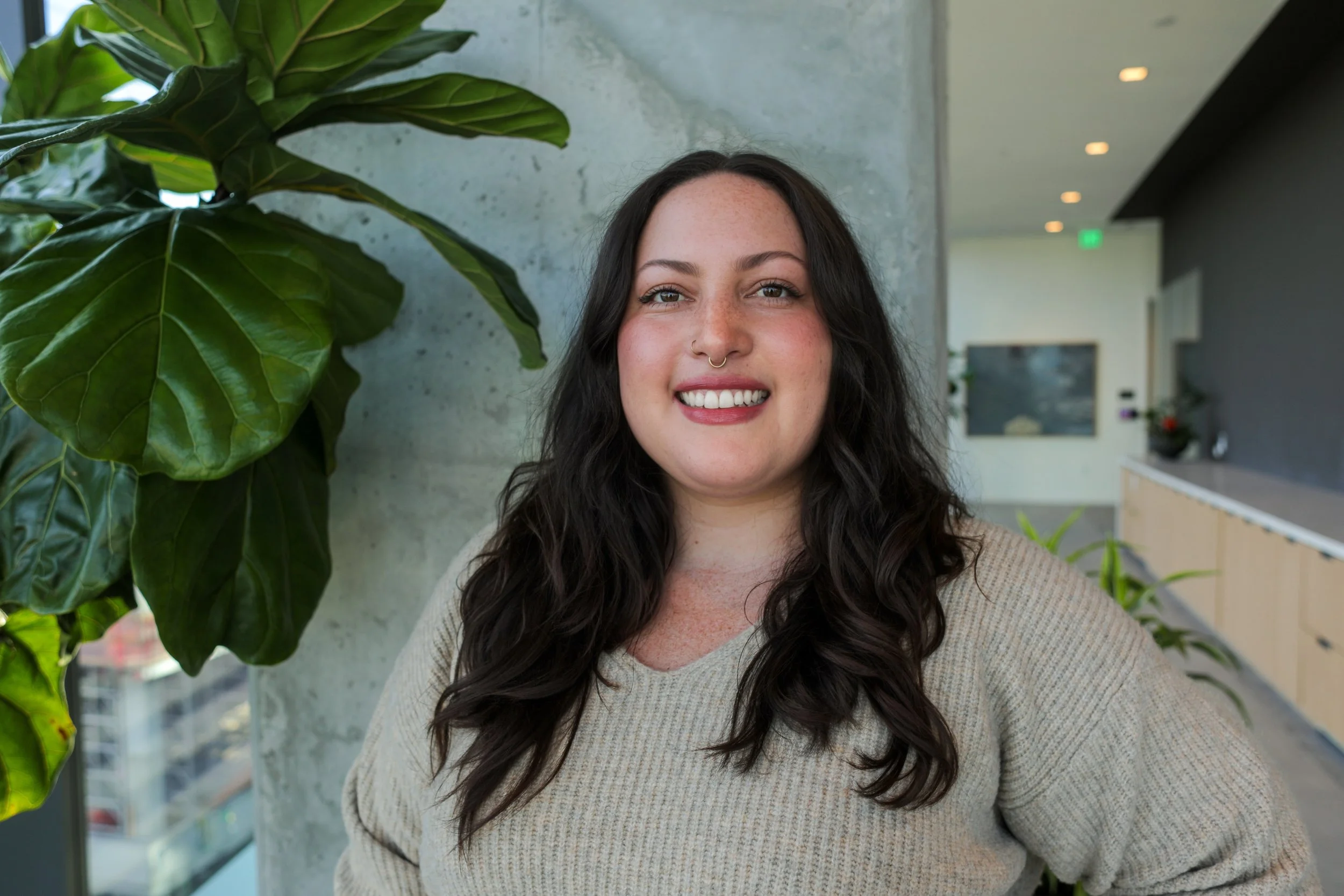 A smiling young woman with dark wavy hair and a septum nose ring, posing indoors next to a large green houseplant, with a city view visible through the window.