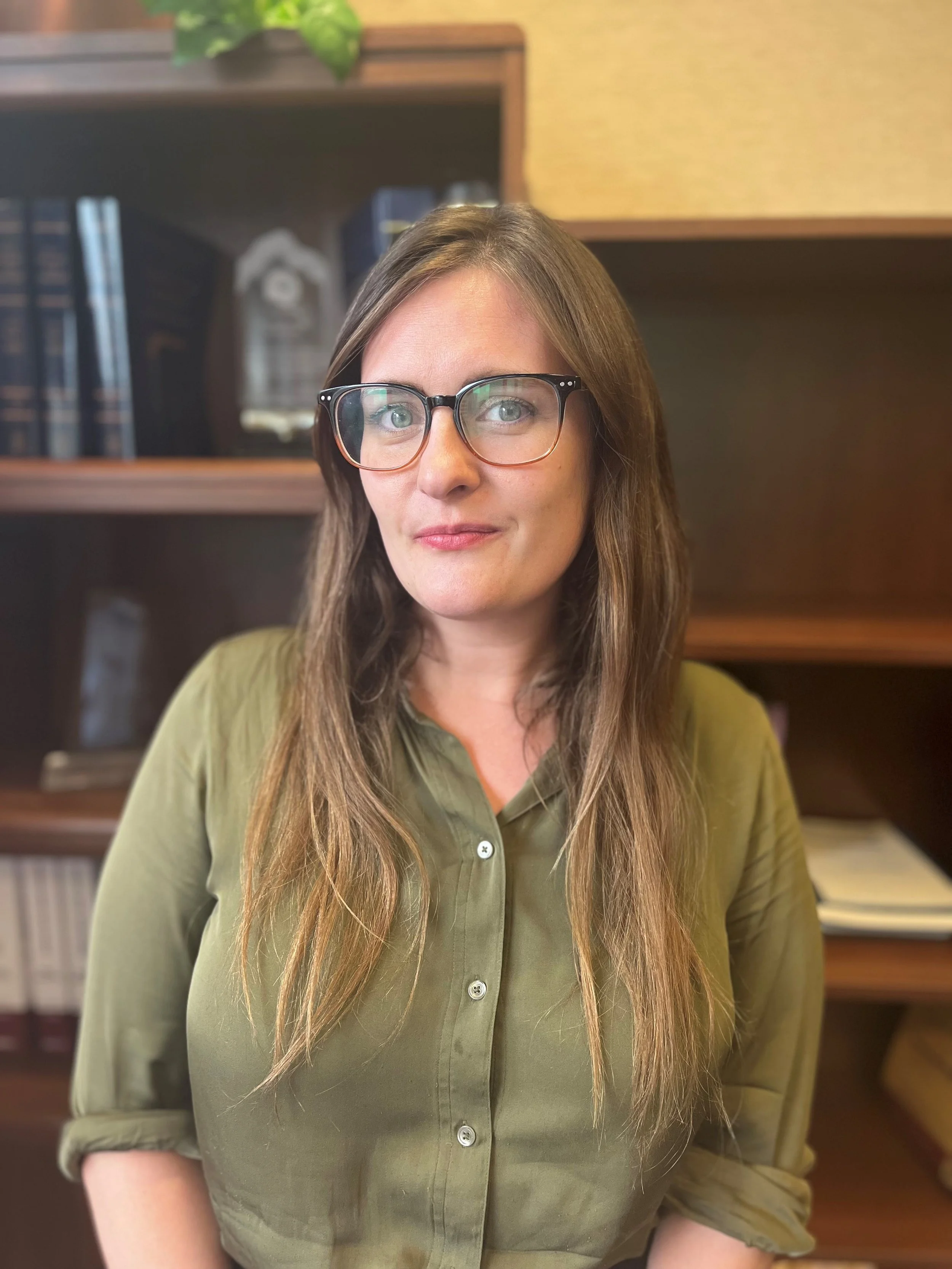 A woman with long brown hair and glasses wearing a green button-up shirt, standing in front of a bookshelf in an indoor setting.