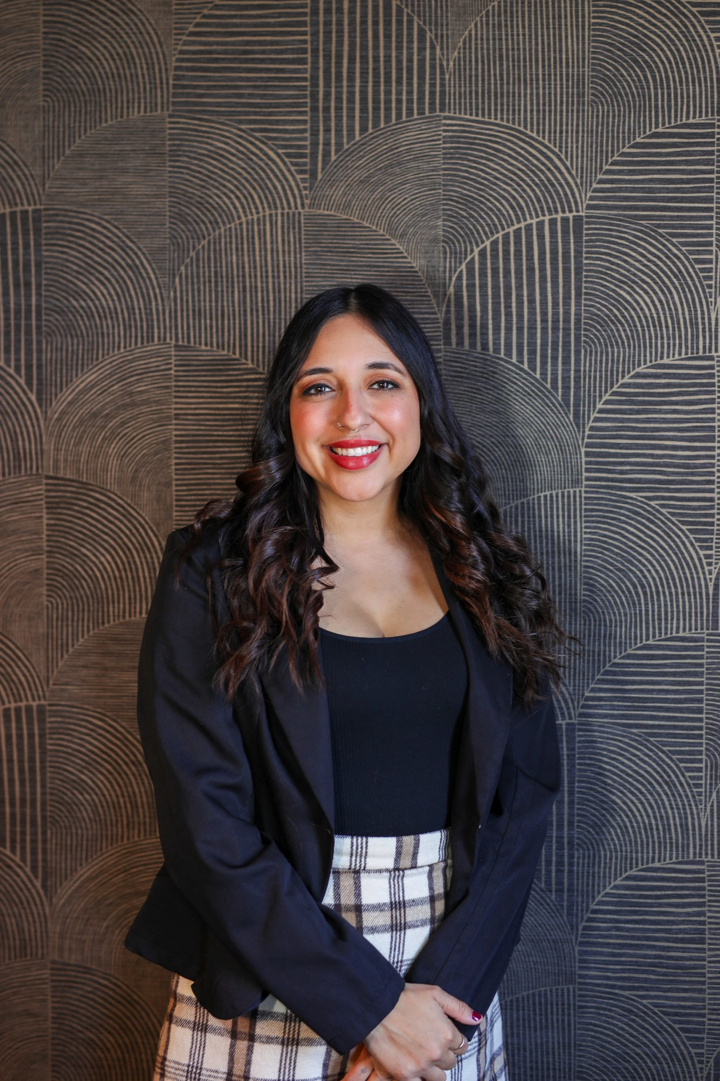 A woman with wavy blonde hair and a nose piercing, wearing a white blouse and a reddish-brown blazer, smiling in front of a green plant wall.