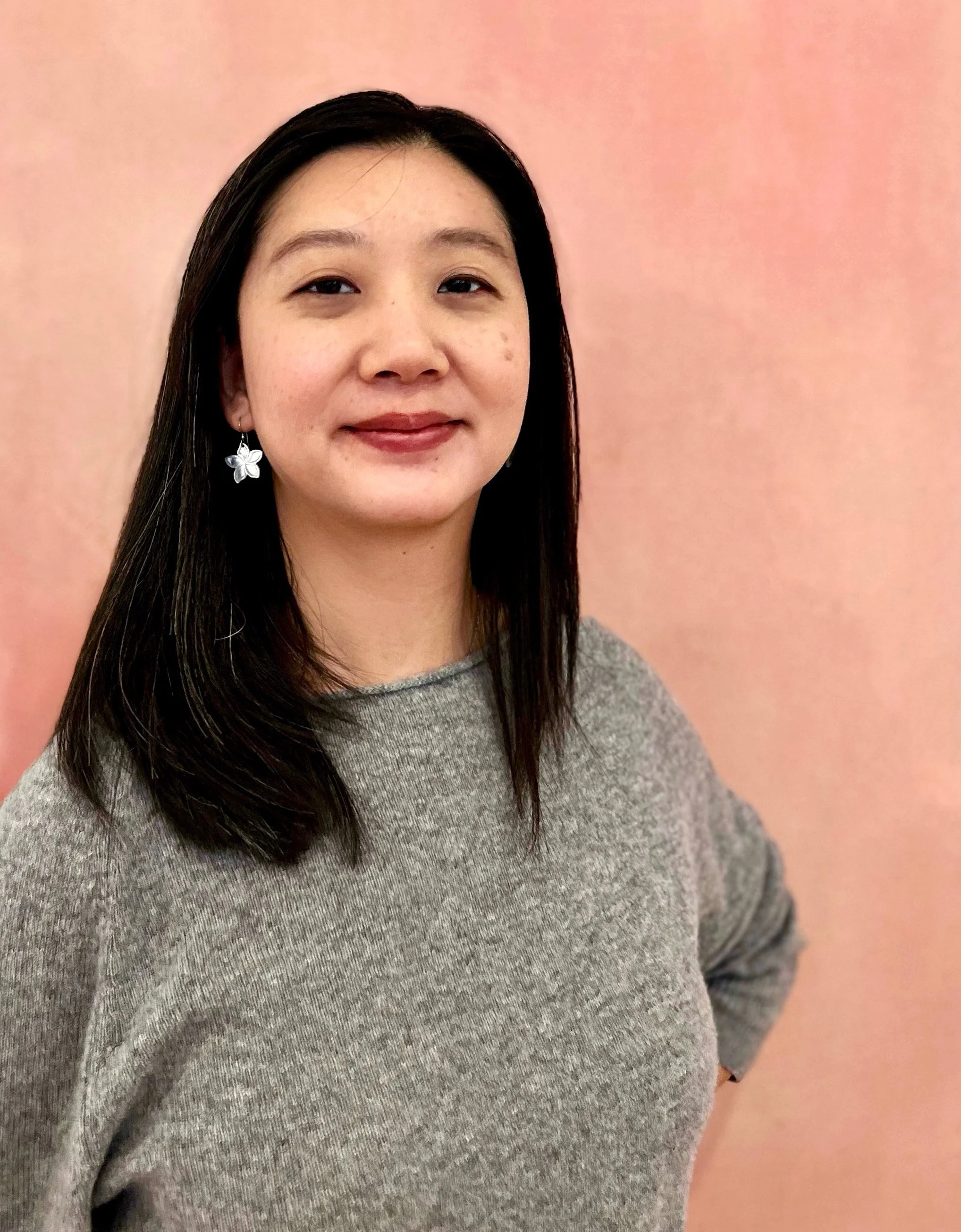 A woman with long black hair wearing a grey sweater and butterfly earrings standing in front of a pink wall.