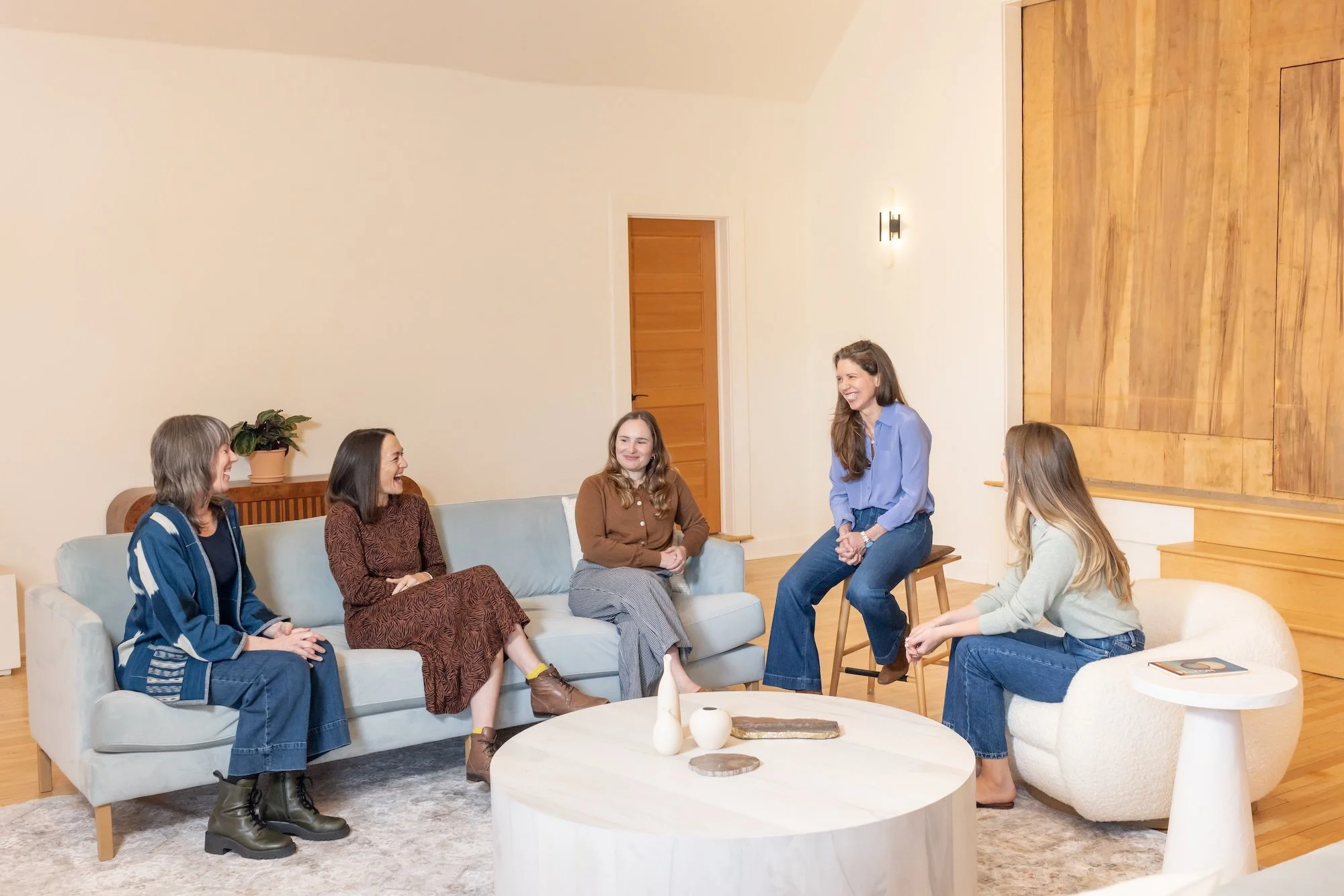 Group of women sitting and talking in a modern living room with light wood decor and neutral furniture.