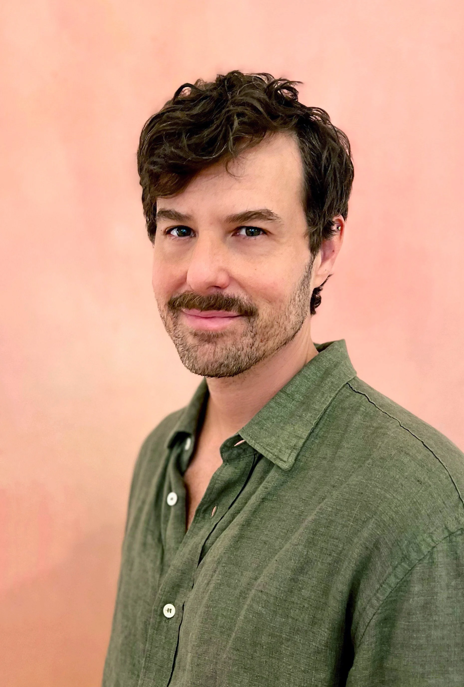 A man with short curly dark hair, blue eyes, and a beard, wearing a green shirt, standing against a pinkish background.