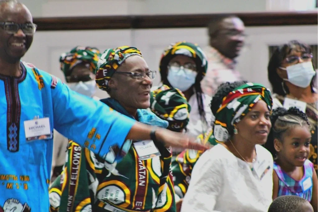 Group of women and children wearing colorful traditional African clothing and head wraps, some wearing face masks, standing indoors at an event.