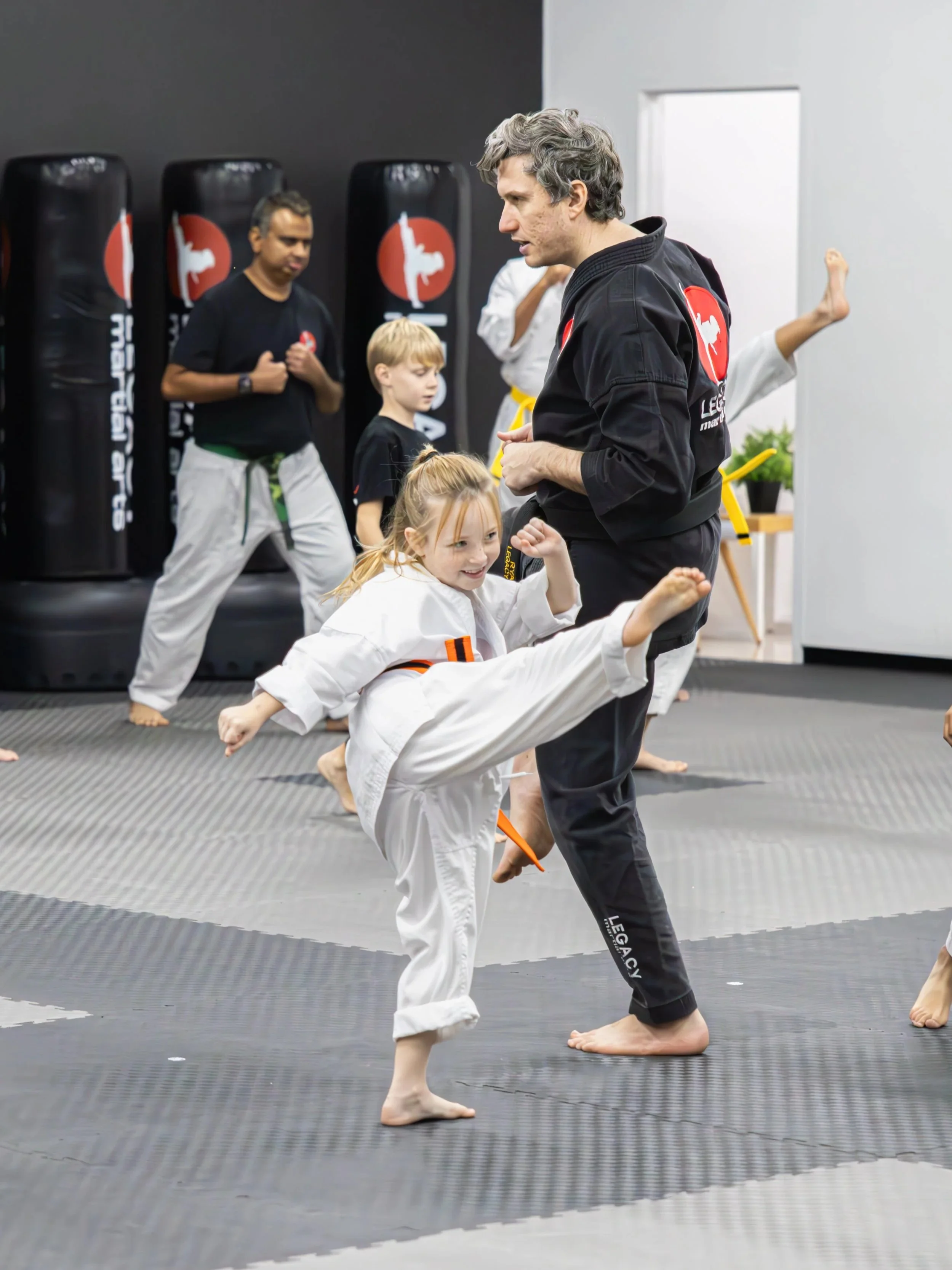 A little girl in a white martial arts uniform in North Lakes, Brisbane, practicing a high kick in a martial arts training room, with a man in a black uniform coaching her. Several other children and instructors are visible in the background.