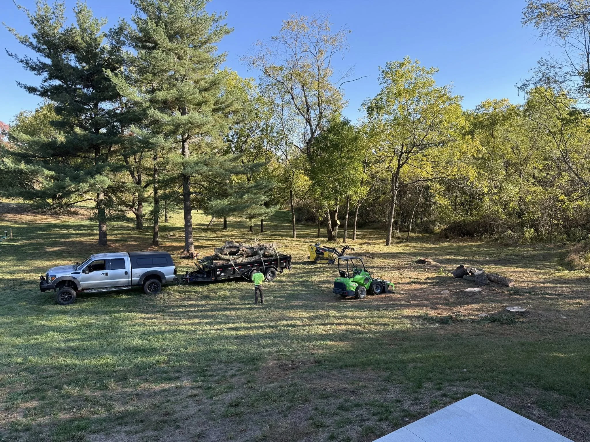 A backyard scene with tall trees and a clear blue sky, including a white truck, a trailer with logs, a green lawn tractor, and two men working near some wood logs.