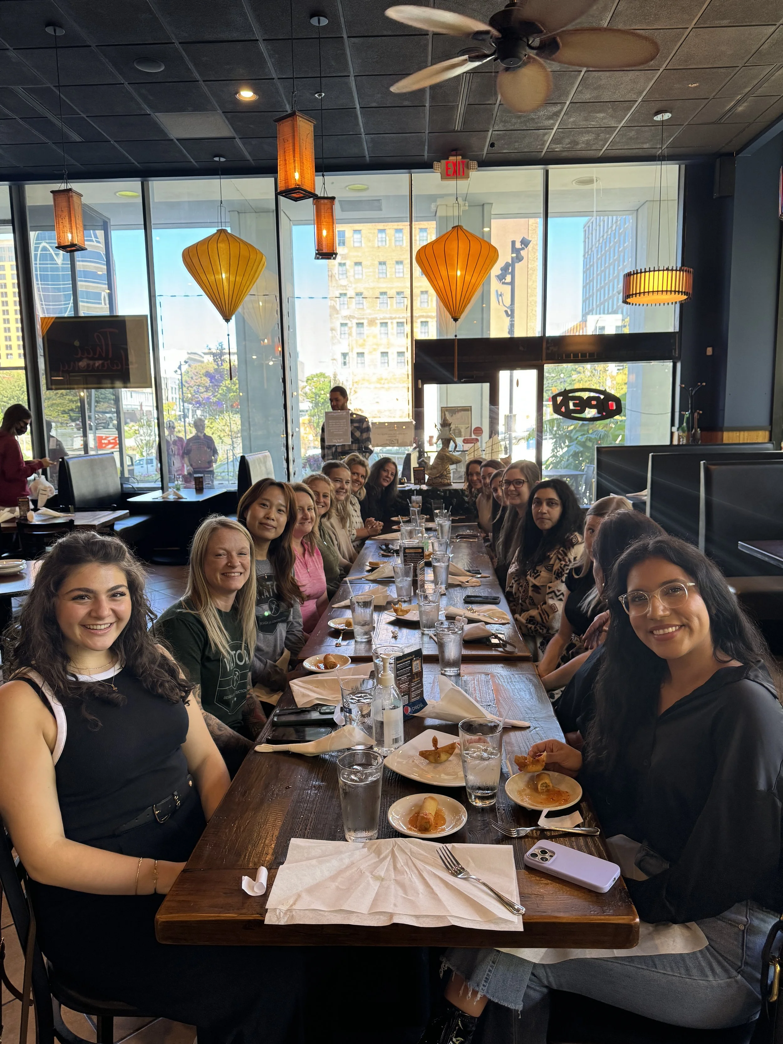 A group of women sitting at a long table in a restaurant, smiling for the photo, with plates of food and glasses of water in front of them, large windows with city buildings in the background, and warm hanging lights.