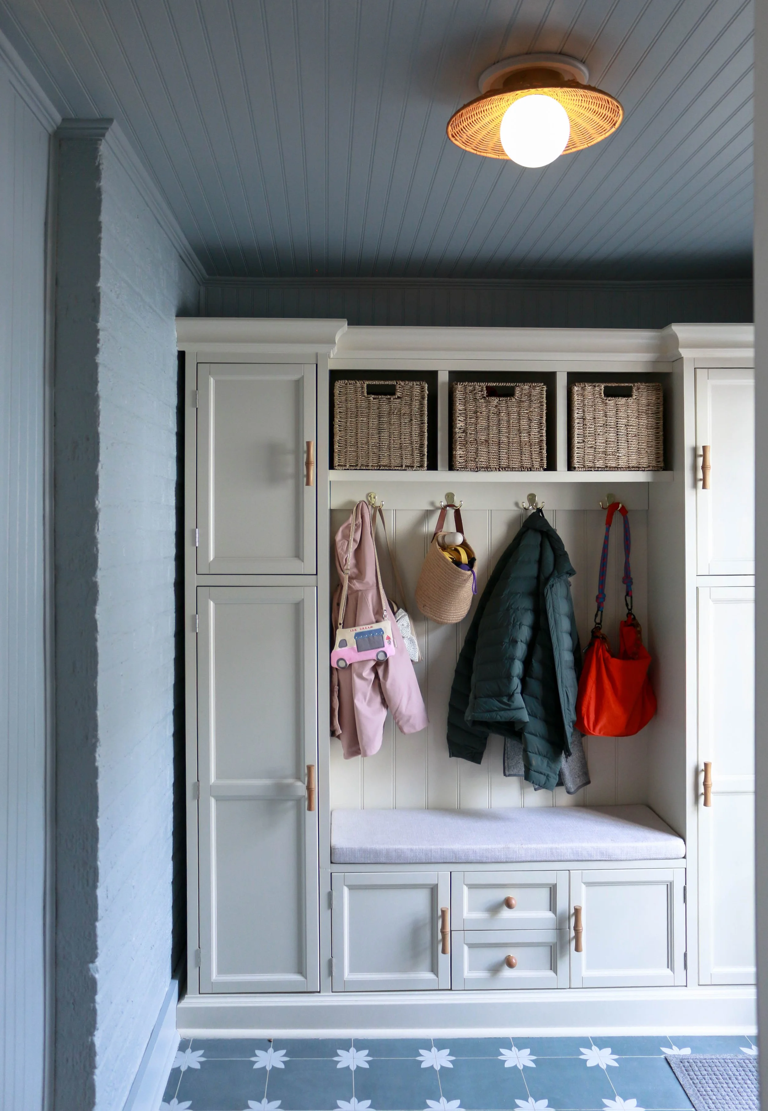 A white hall tree with storage cabinets and hooks, holding kids' coats, bags, and toys, with woven baskets on top and a cushioned seat.