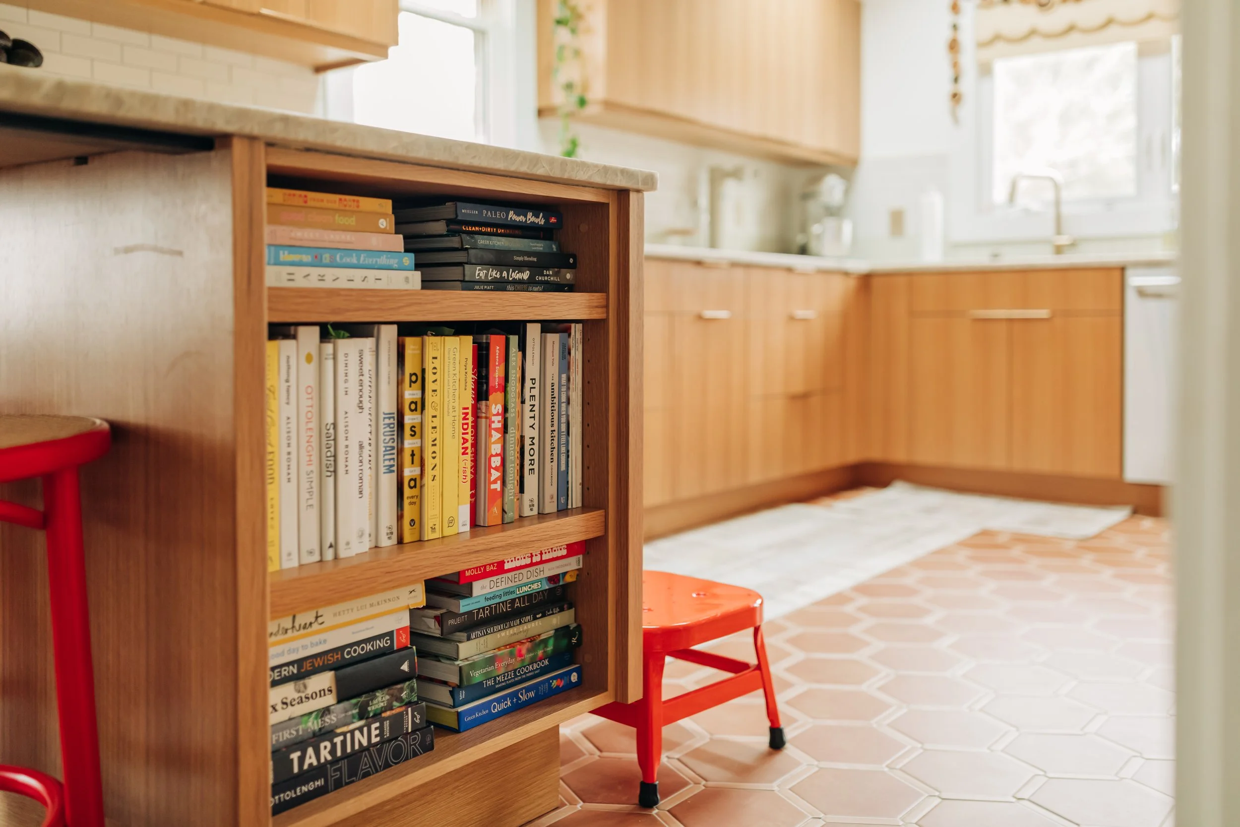 A kitchen with wooden cabinets and beige tiled flooring. A bookshelf filled with cookbooks and food-related books stands in the foreground. There is a small red chair and a white rug on the floor near the cabinets. A window is visible in the background.