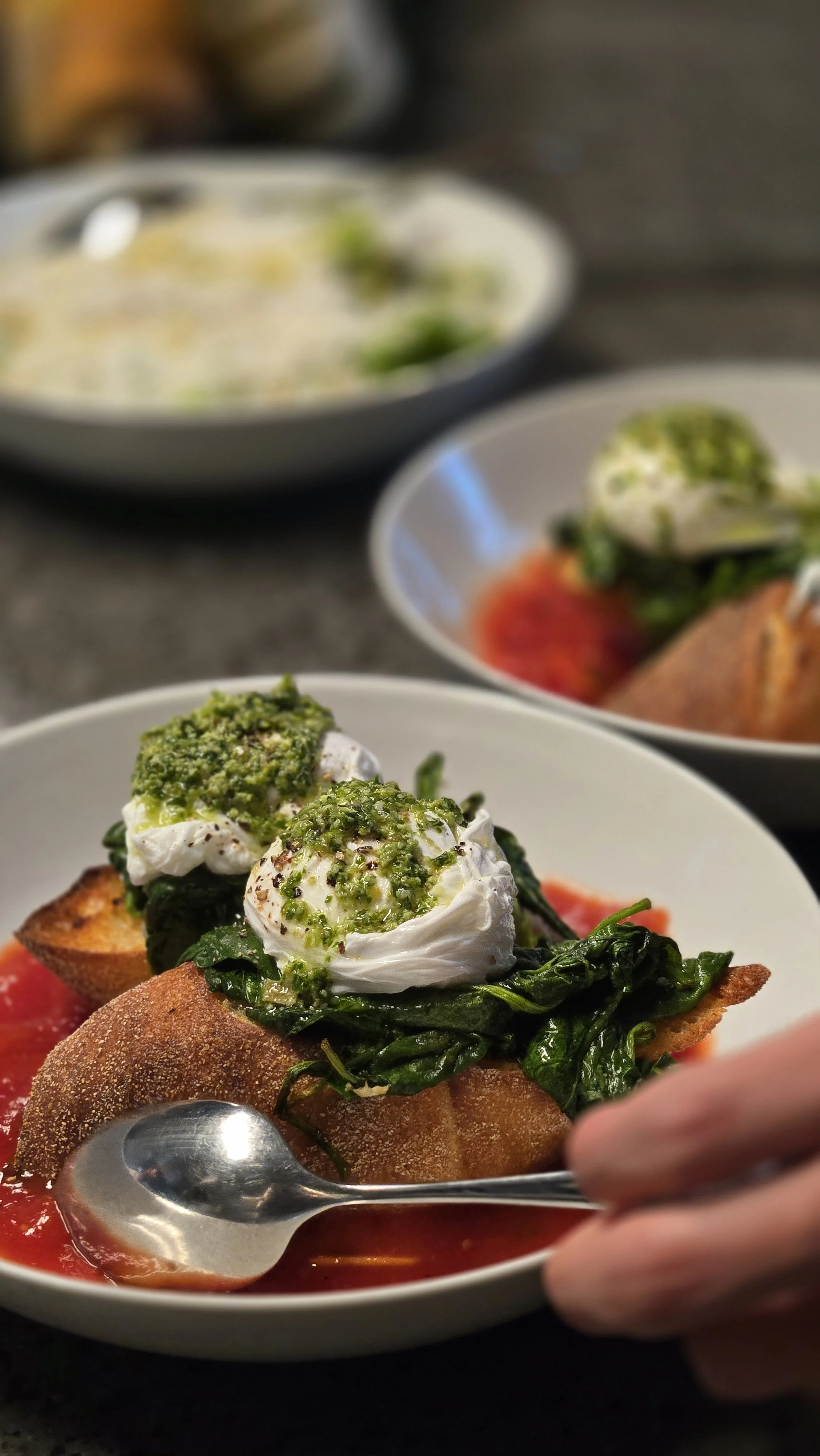 Close-up of a plate with an appetizer topped with a poached egg, spinach, green pesto, and a crispy breadstick, with a spoon partially in the frame and blurred dishes in the background.