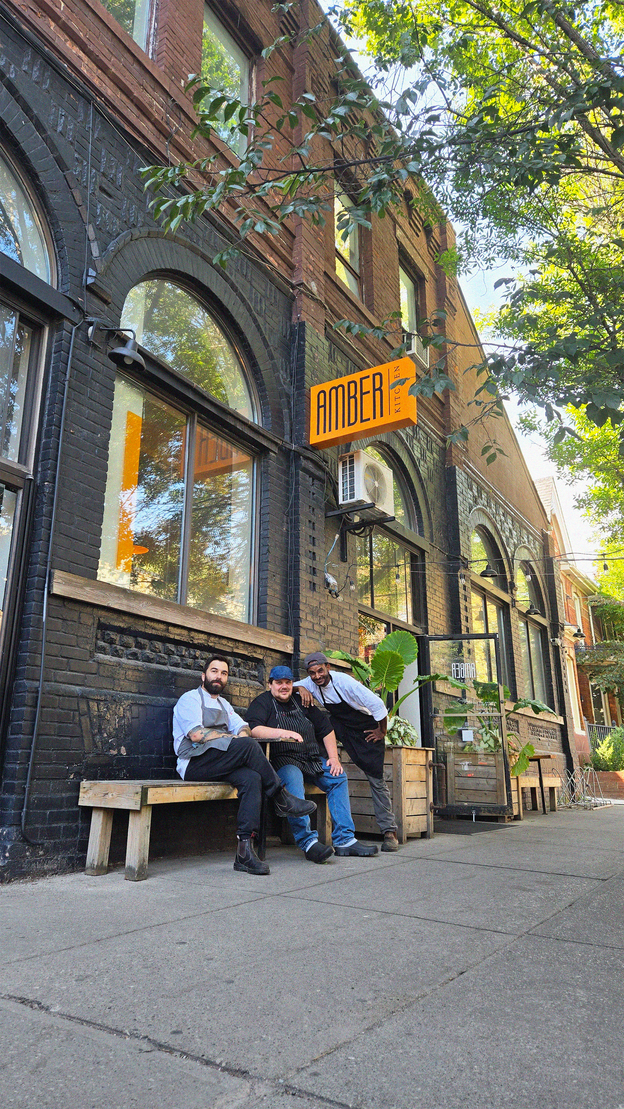 Three chefs sitting on a wooden bench outside a brick restaurant called Amber Kitchen. The restaurant has large arched windows, black brick exterior, and a sign with orange background and black text. The chefs are smiling and posing for the photo, with greenery and trees visible in the background.
