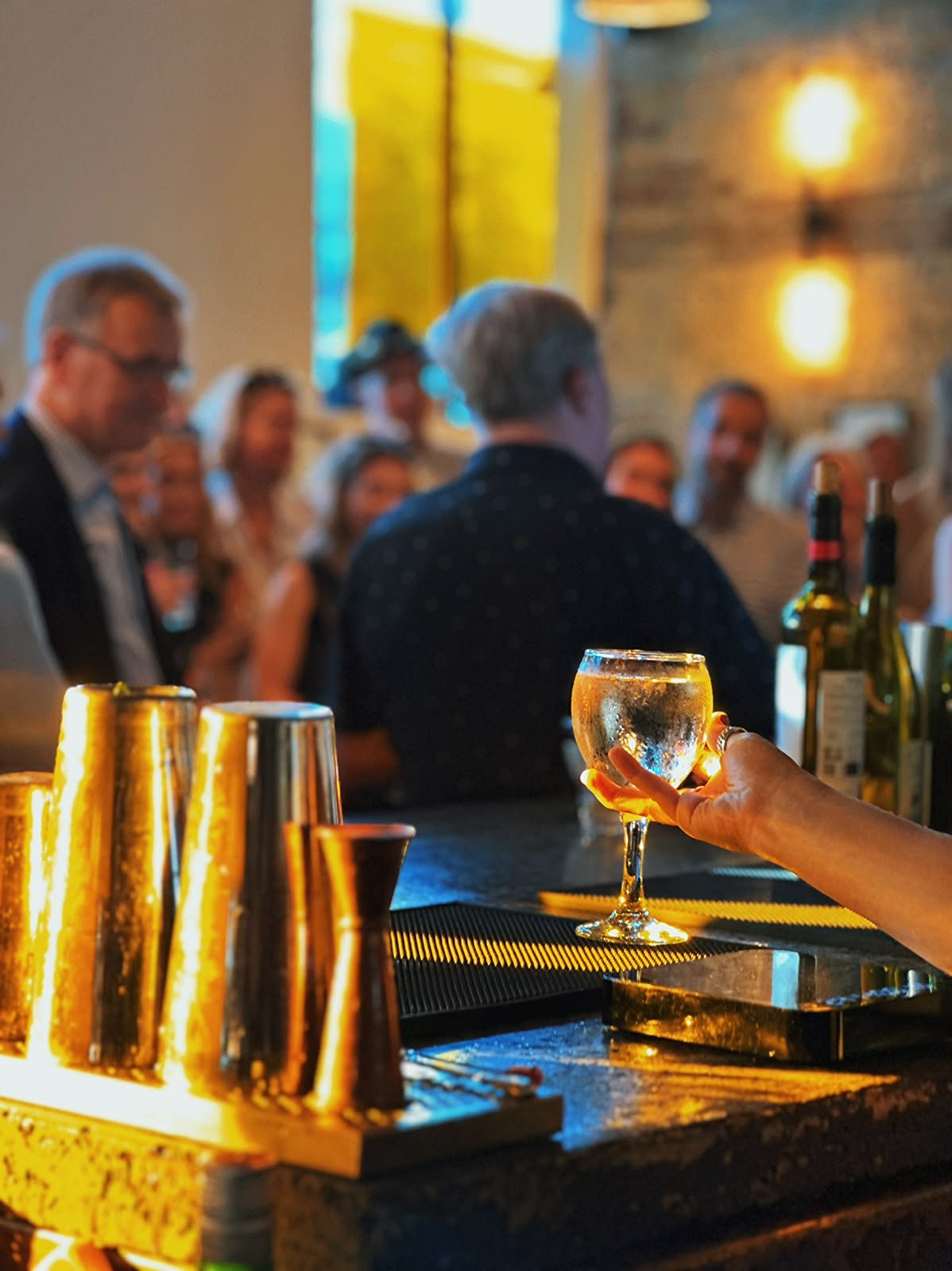 Person holding a glass of white wine at a bar with a group of people in the background, bottles of wine and barware on the counter, warm lighting in a lively bar or restaurant setting.
