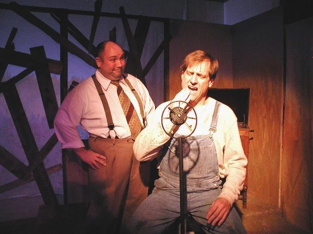 Two men in a recording studio, one singing into a vintage microphone and the other observing.